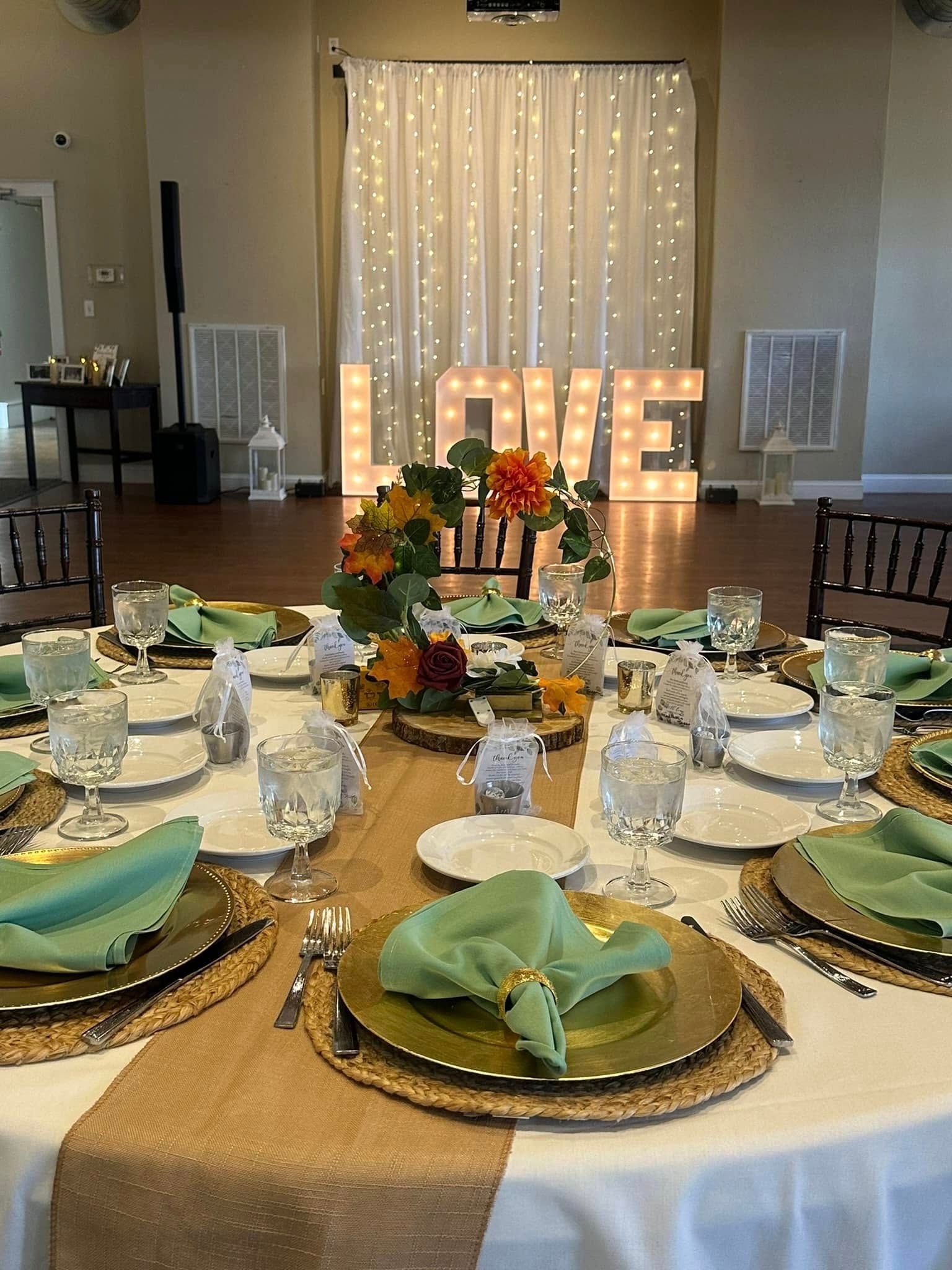 A table set for a wedding reception with plates , glasses , napkins and a love sign in the background.