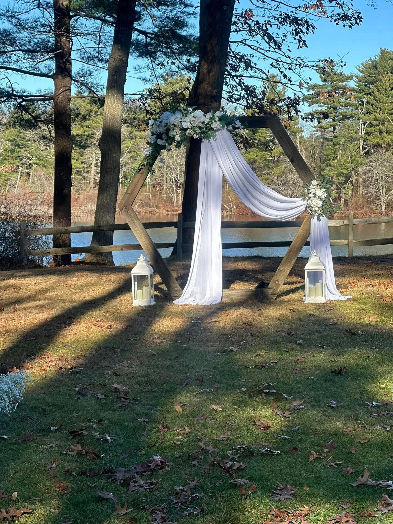 A wooden arch decorated with white flowers and lanterns in front of a lake.