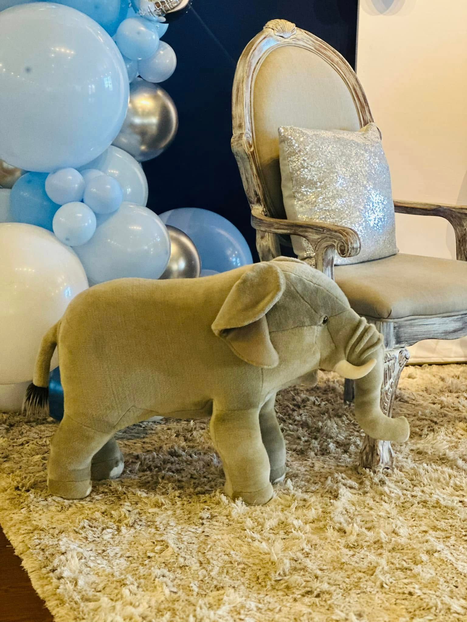 A stuffed elephant is sitting on a rug in front of a chair and balloons.