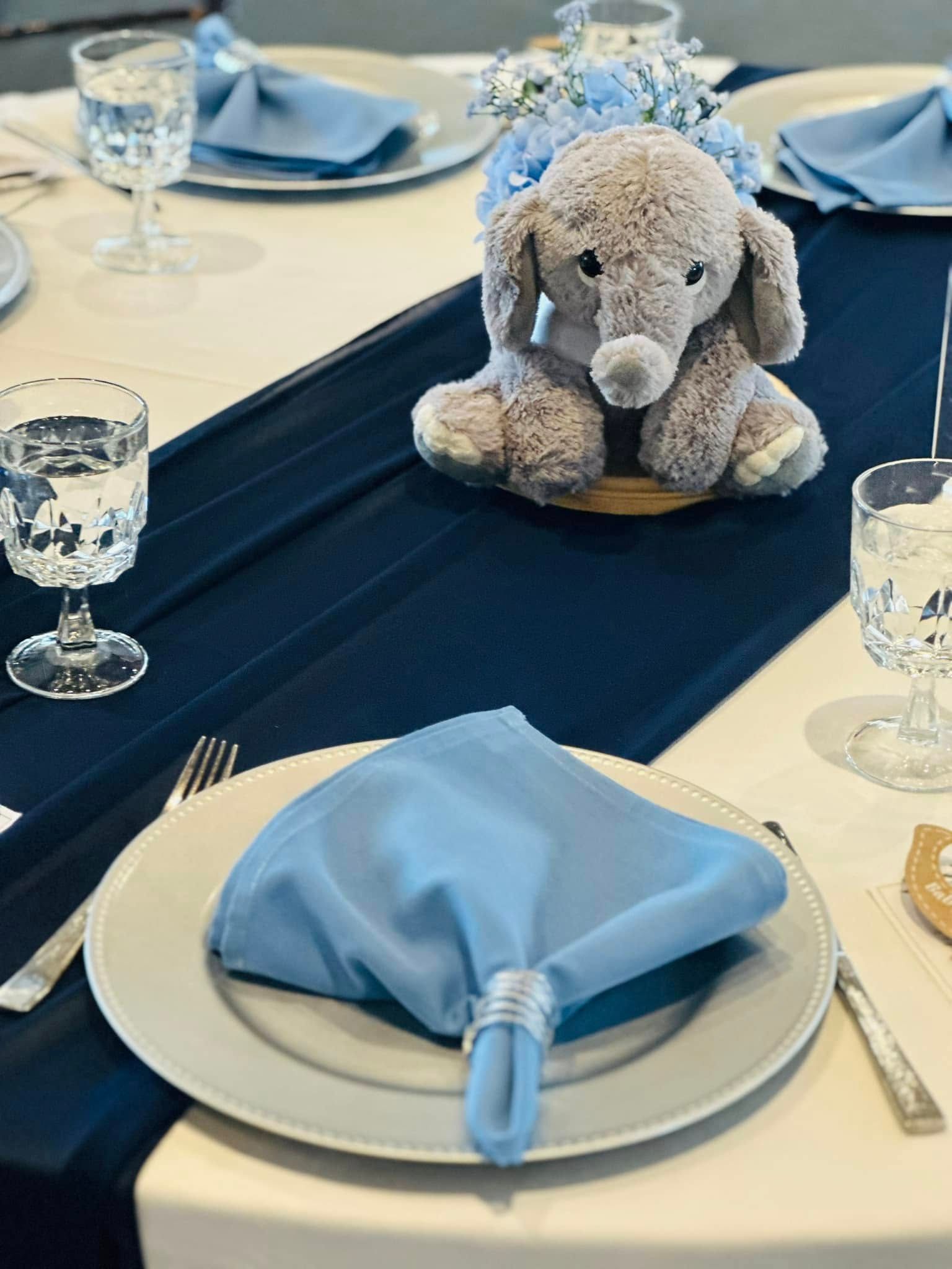 A stuffed elephant is sitting on a table with plates and glasses.