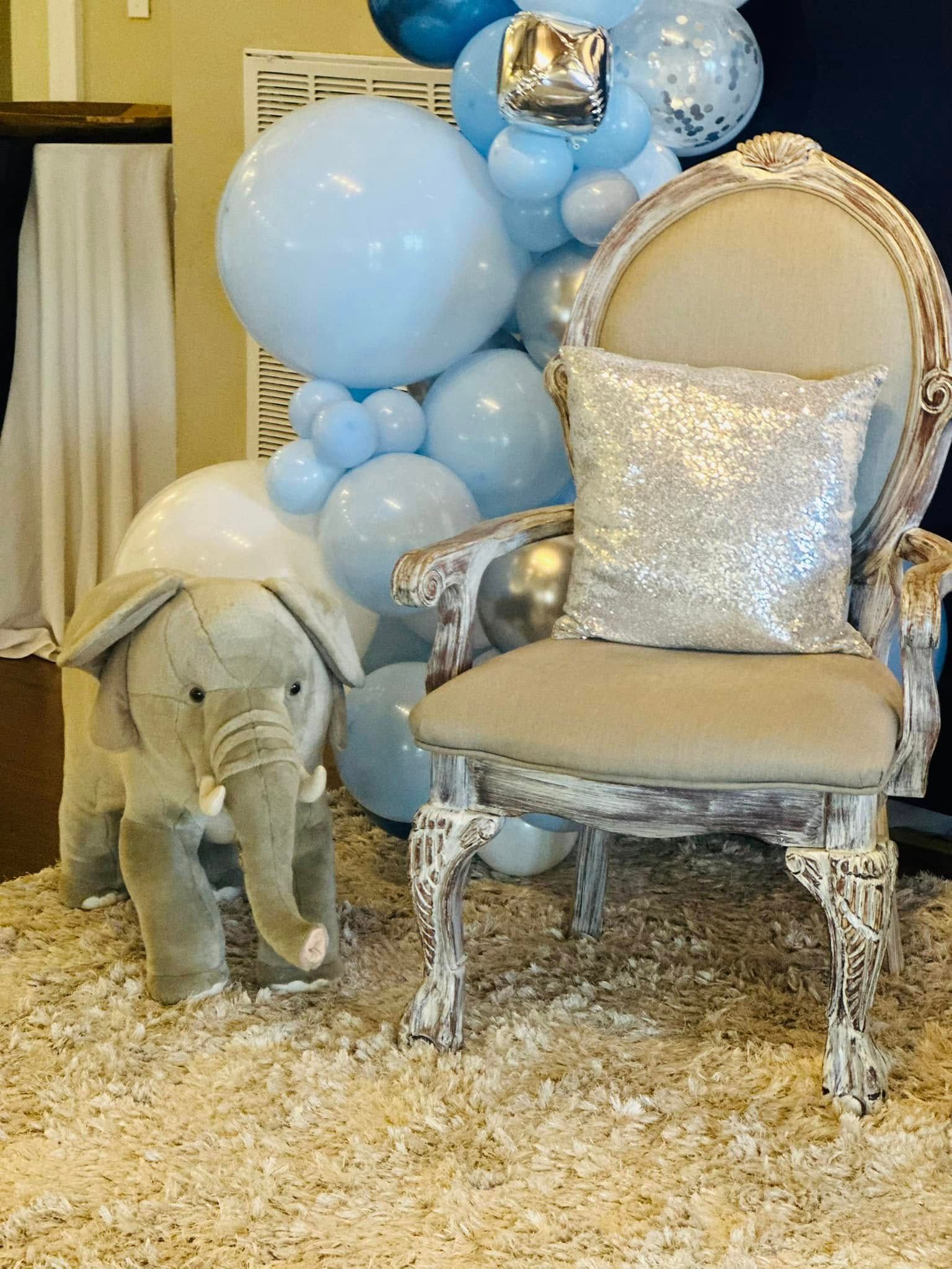 A stuffed elephant is sitting next to a chair with balloons in the background.