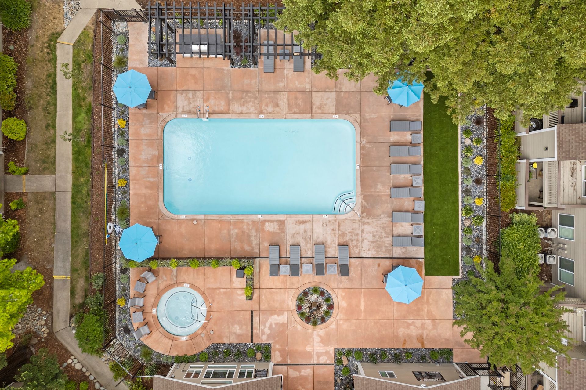 Aerial view of a pool deck with a large rectangular pool, hot tub, lounge chairs, and blue umbrellas.