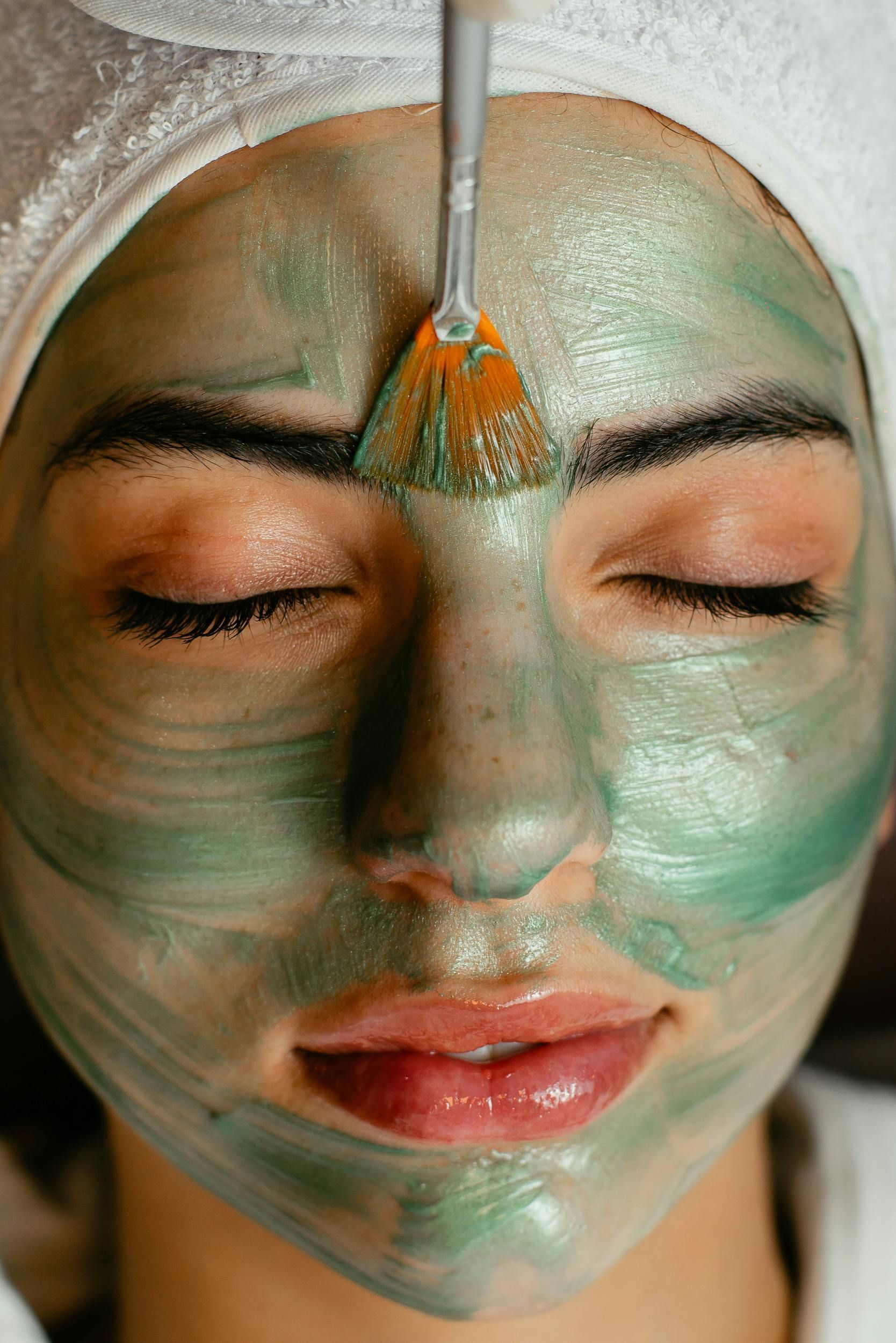 A woman is getting a green facial mask on her face.