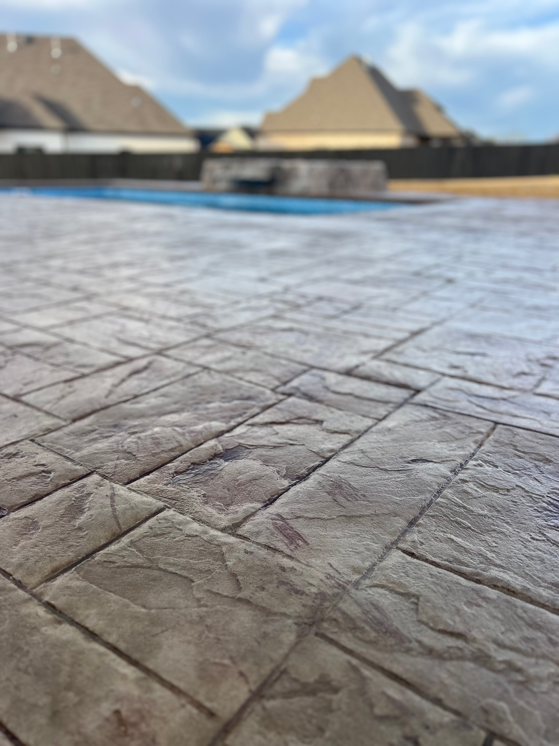 Stamped concrete patio with a textured stone pattern, blurred view of a pool and houses in the background.