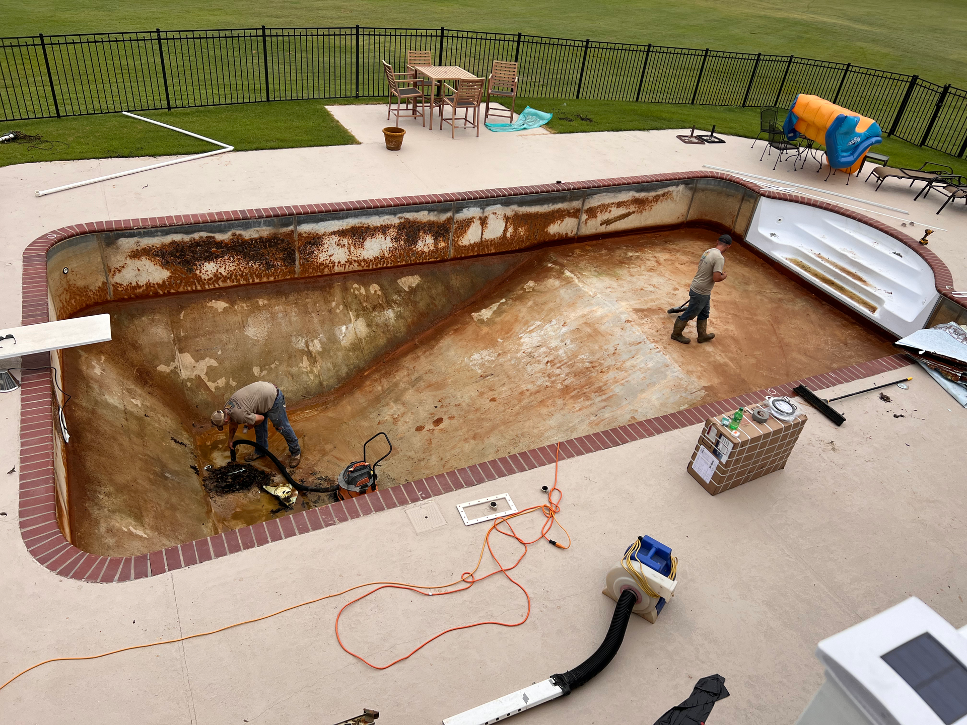 Two workers cleaning a drained, rusty swimming pool. Concrete patio, lawn, and black fence surround.