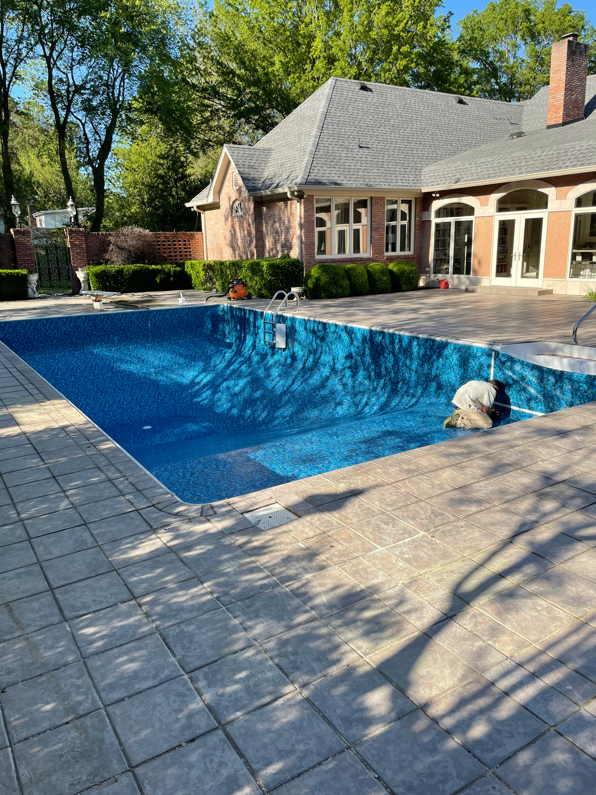 Empty blue pool surrounded by paving stones in front of a brick house with a dark roof.