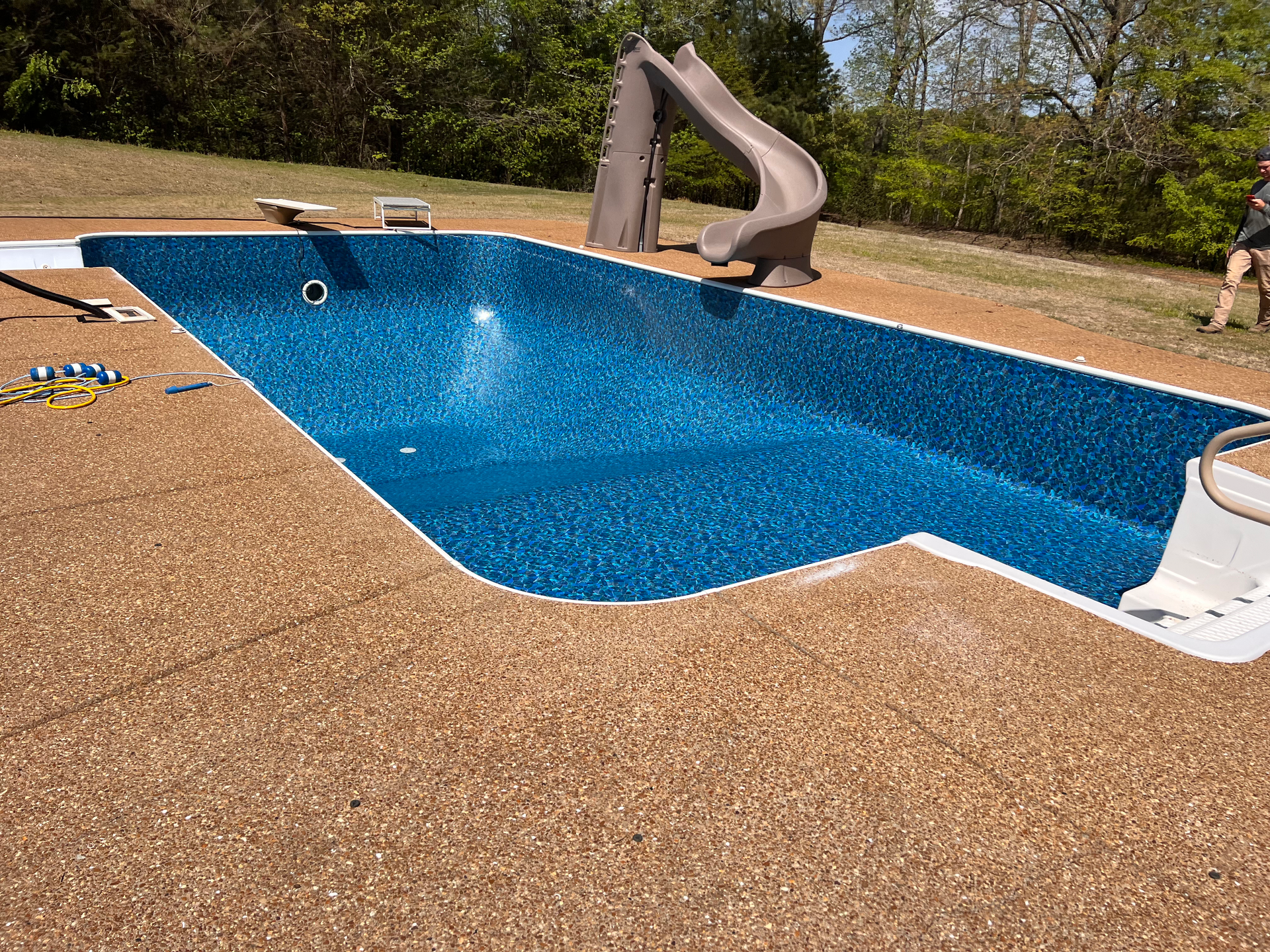 Blue-lined rectangular swimming pool with brown concrete edging, a brown slide, and green grass in background.