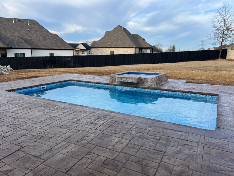 Rectangular swimming pool and small hot tub in a backyard with stamped concrete. Brown fence and houses in the background.