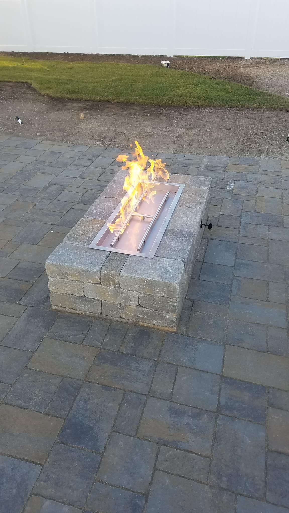 Fire pit with flames, made of gray stone, on a patio. Green grass and white wall in the background.