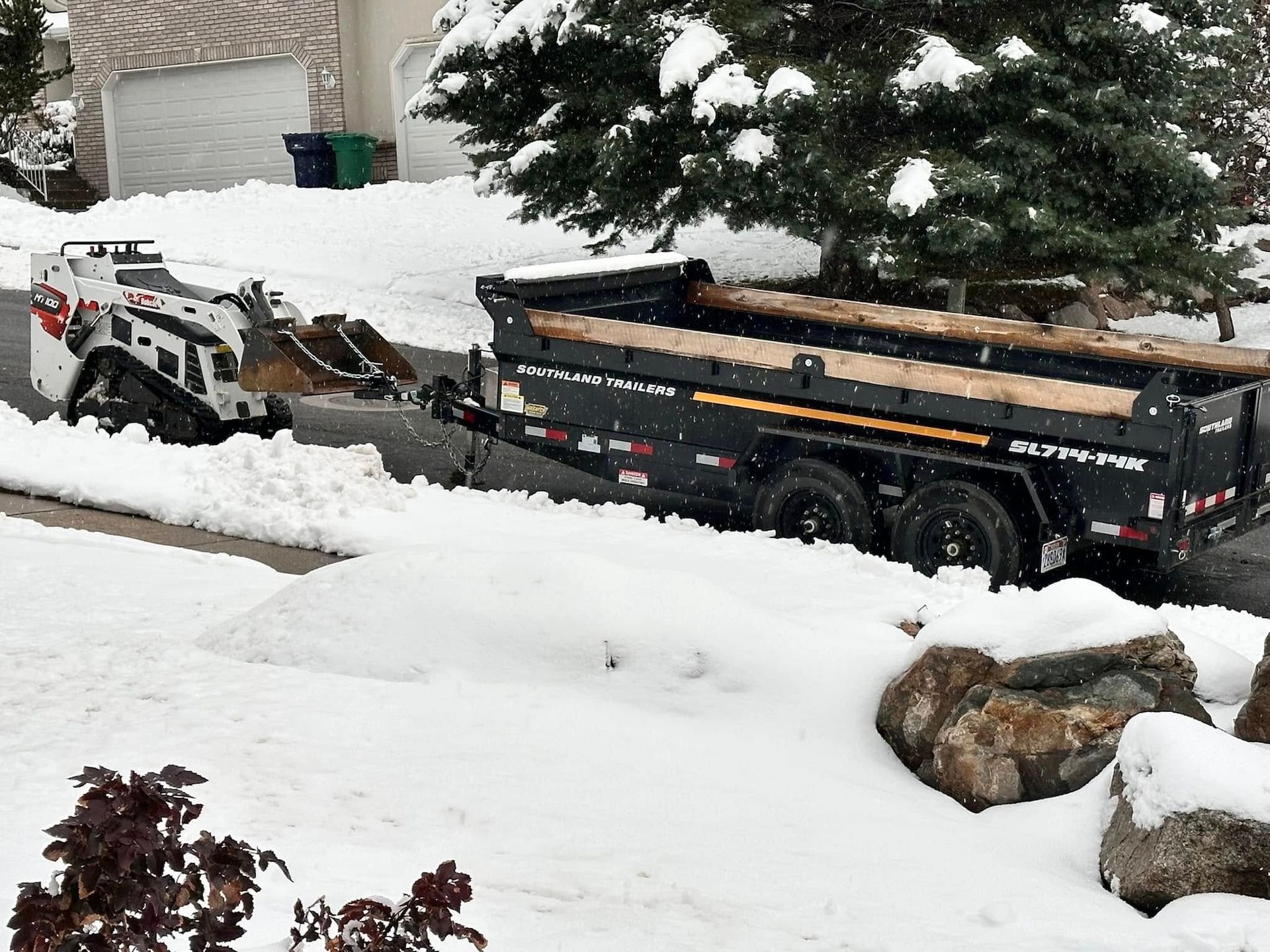 A small tracked skid steer loader clearing snow, loading it into a black dump trailer in a snowy suburban driveway.