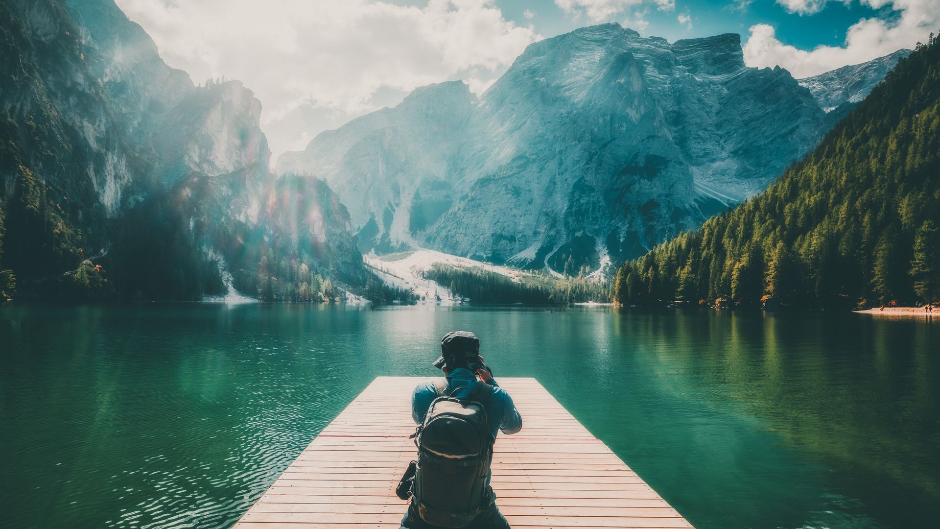 A man is sitting on a dock overlooking a lake with mountains in the background.