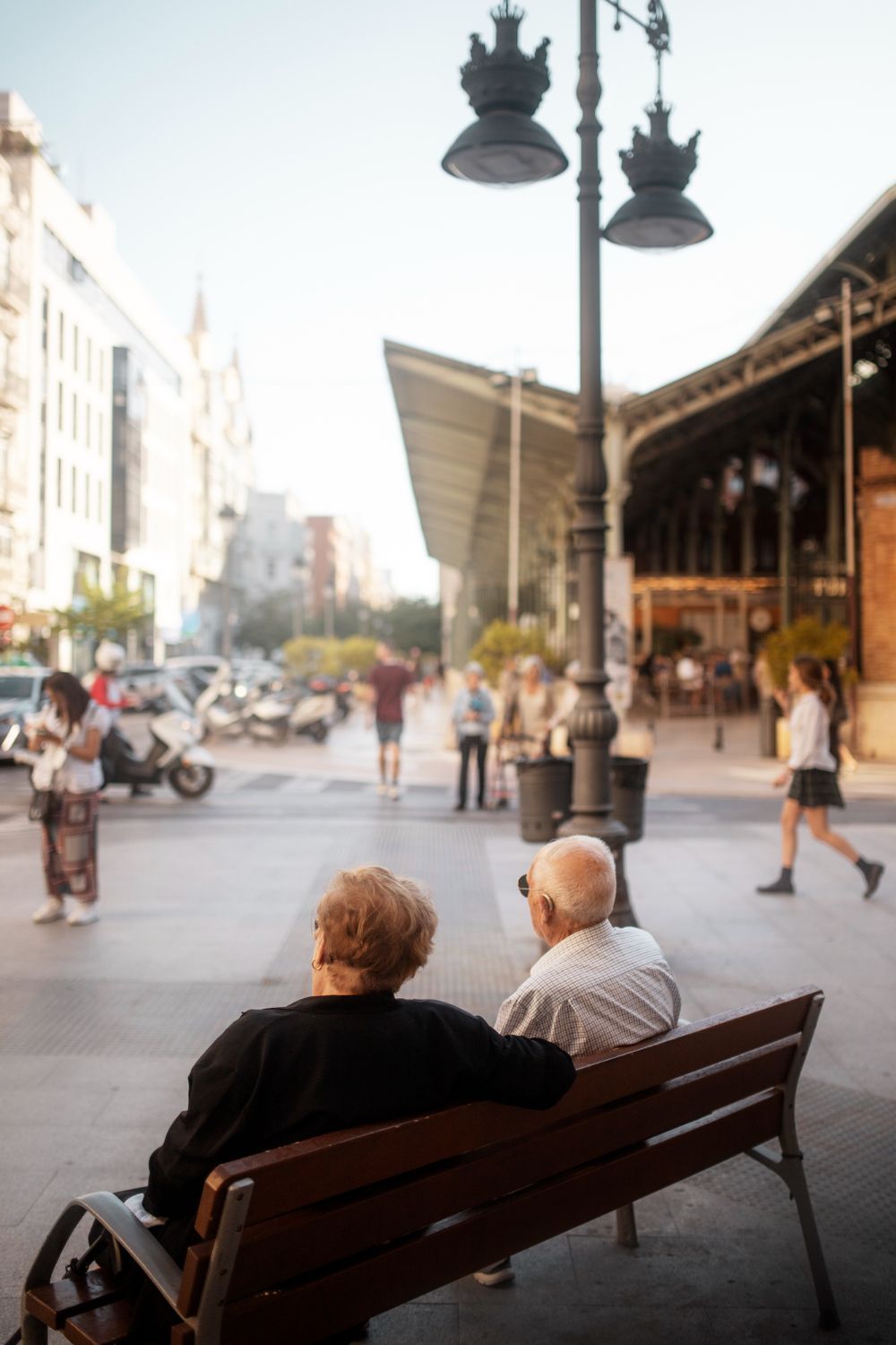 Elderly couple sitting on a bench, people and traffic in the background, city street scene.