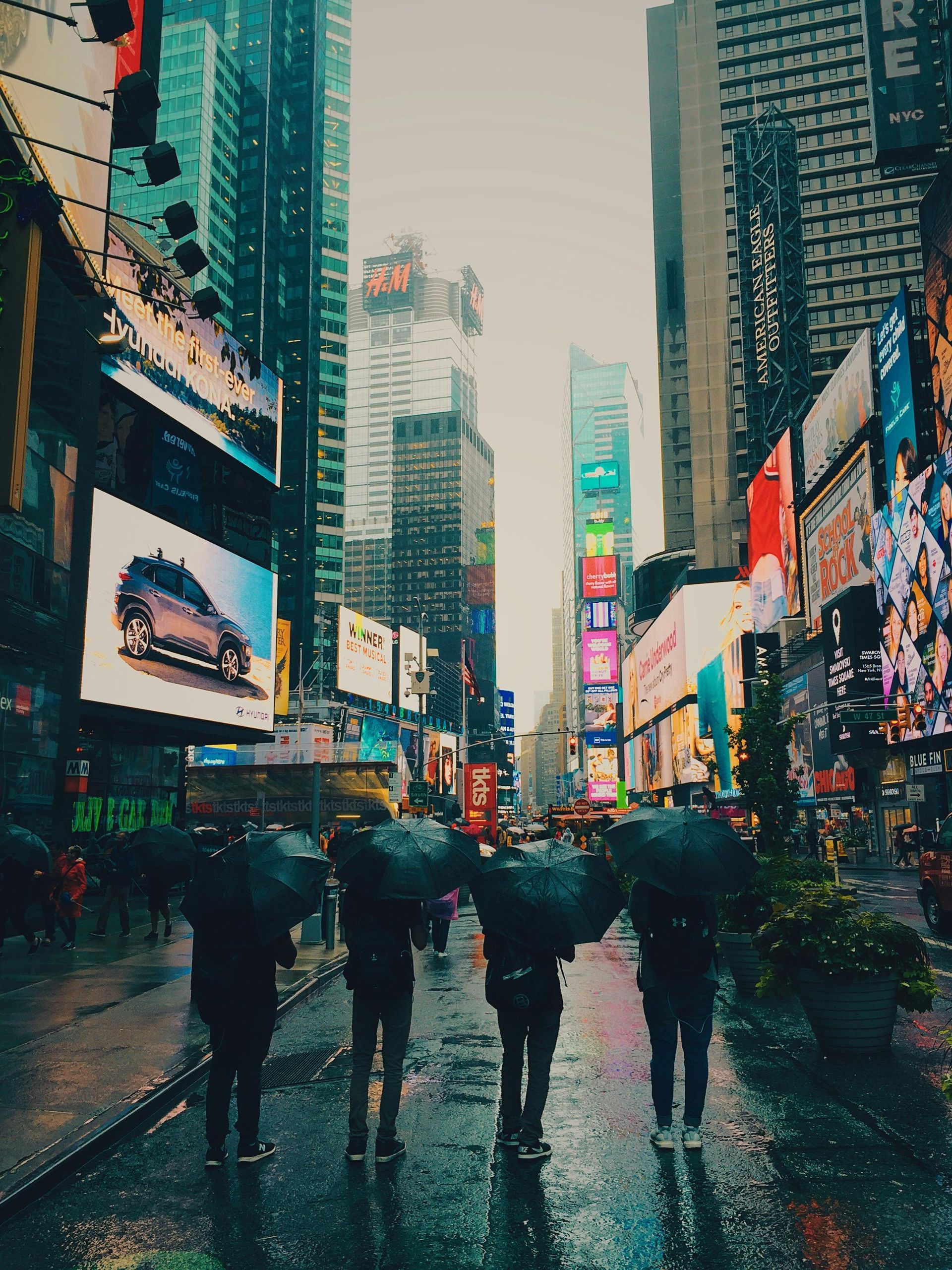 Four people with umbrellas walking in Times Square, New York City, in the rain, surrounded by bright signs.