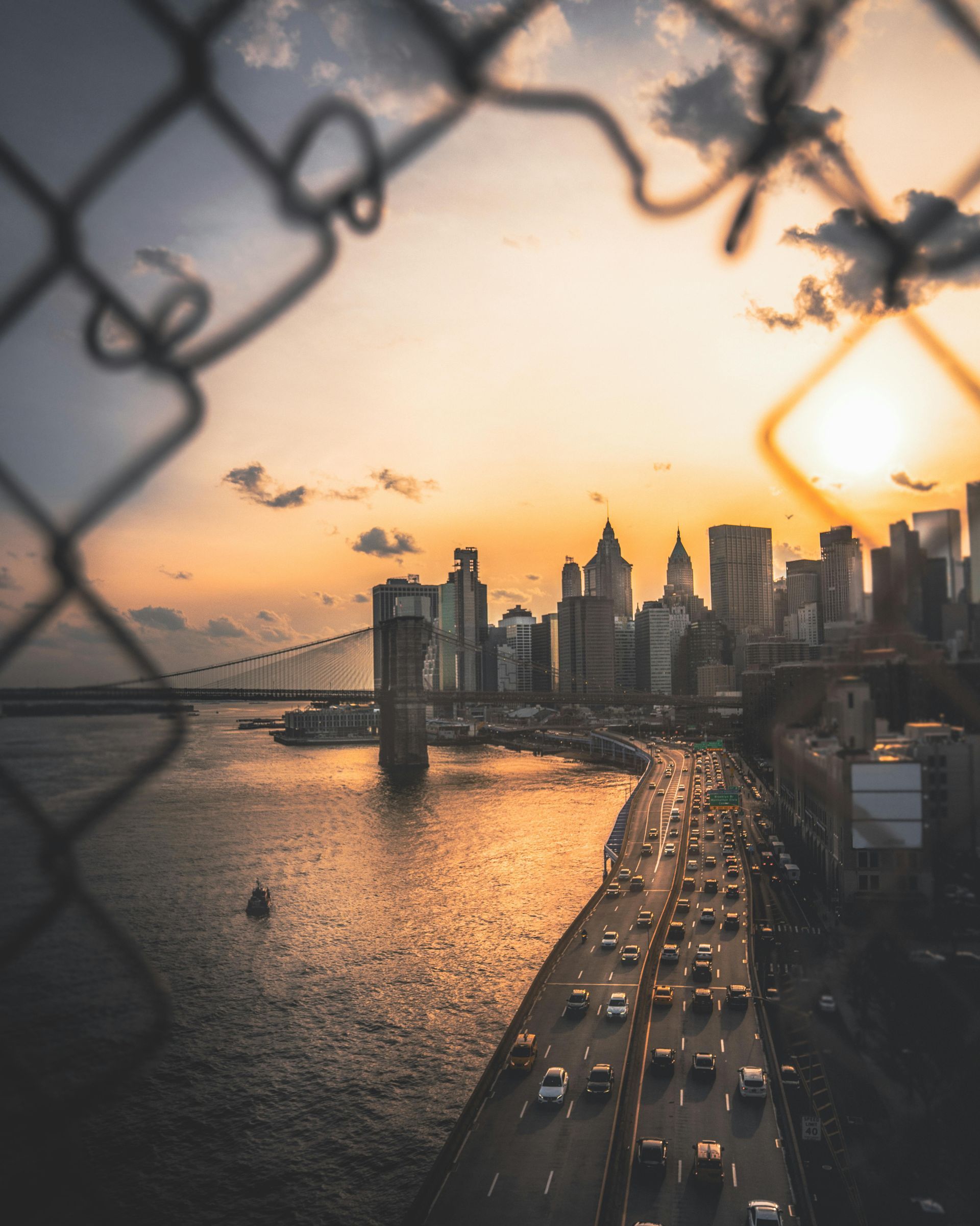 Sunset over New York City skyline, framed by a chain link fence, traffic on highway.