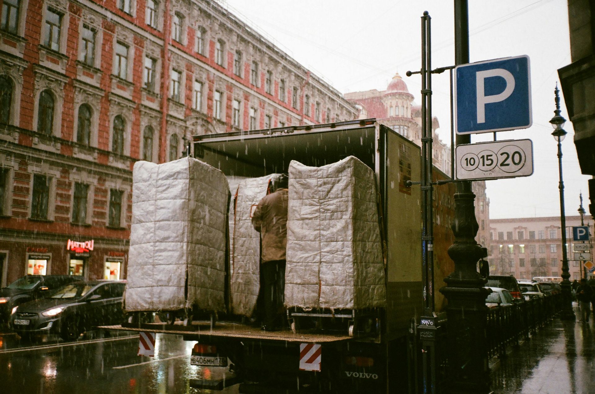 Truck unloading wrapped objects on a rainy city street, parking sign nearby.