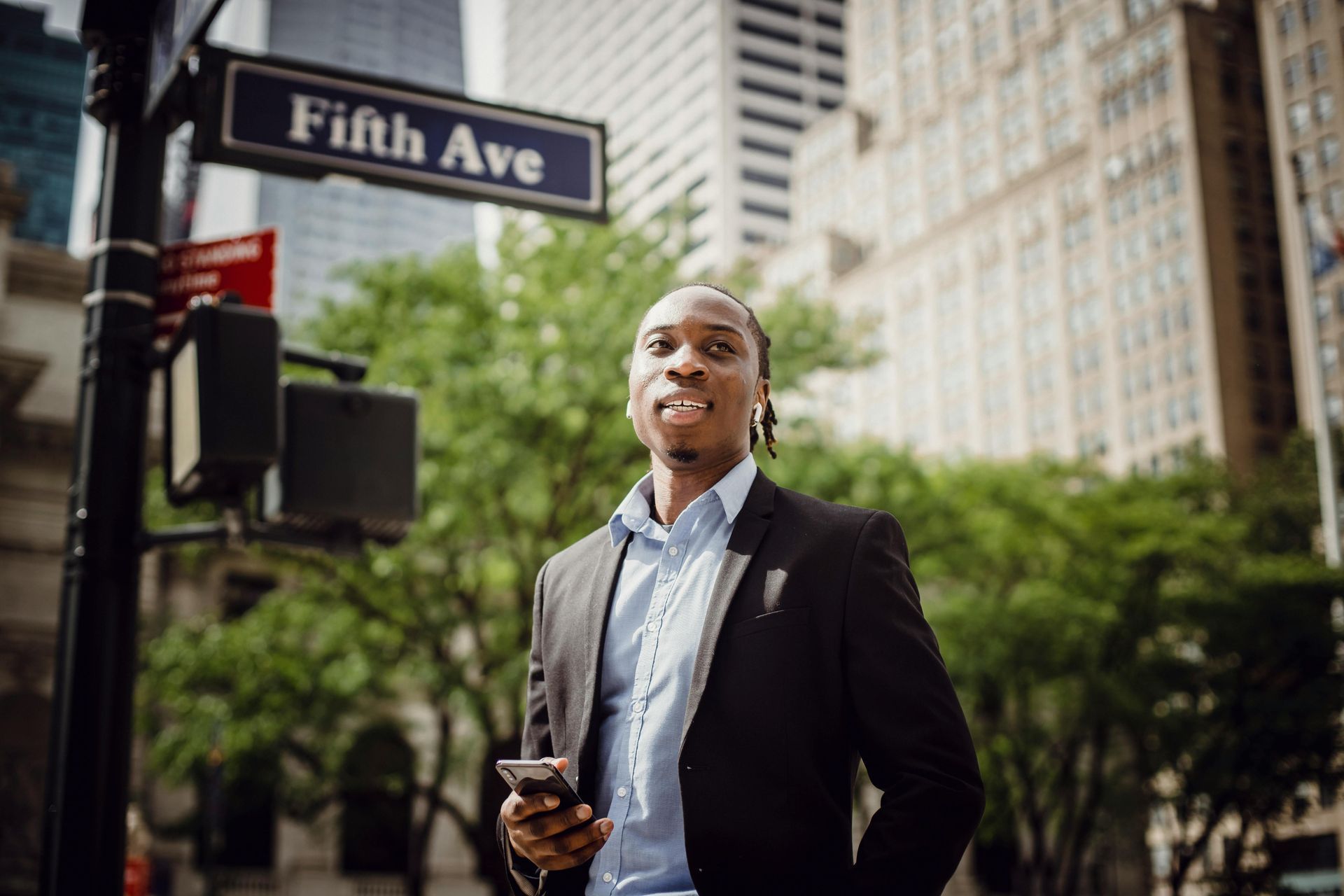 Man on Fifth Ave. holding phone, wearing blazer and button-down. Trees and tall buildings in background.