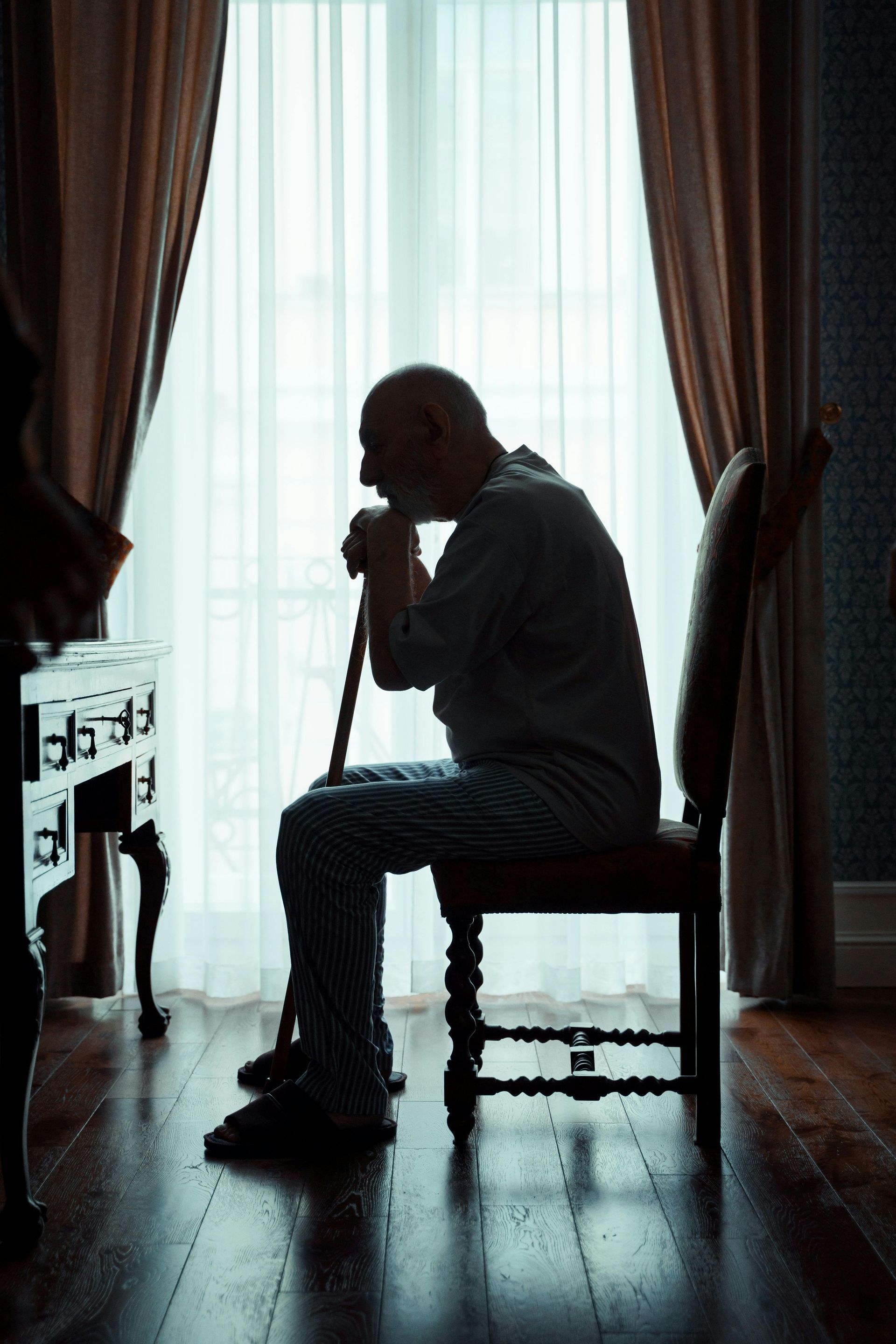Elderly man in silhouette sits, leaning on cane, near a window with curtains.