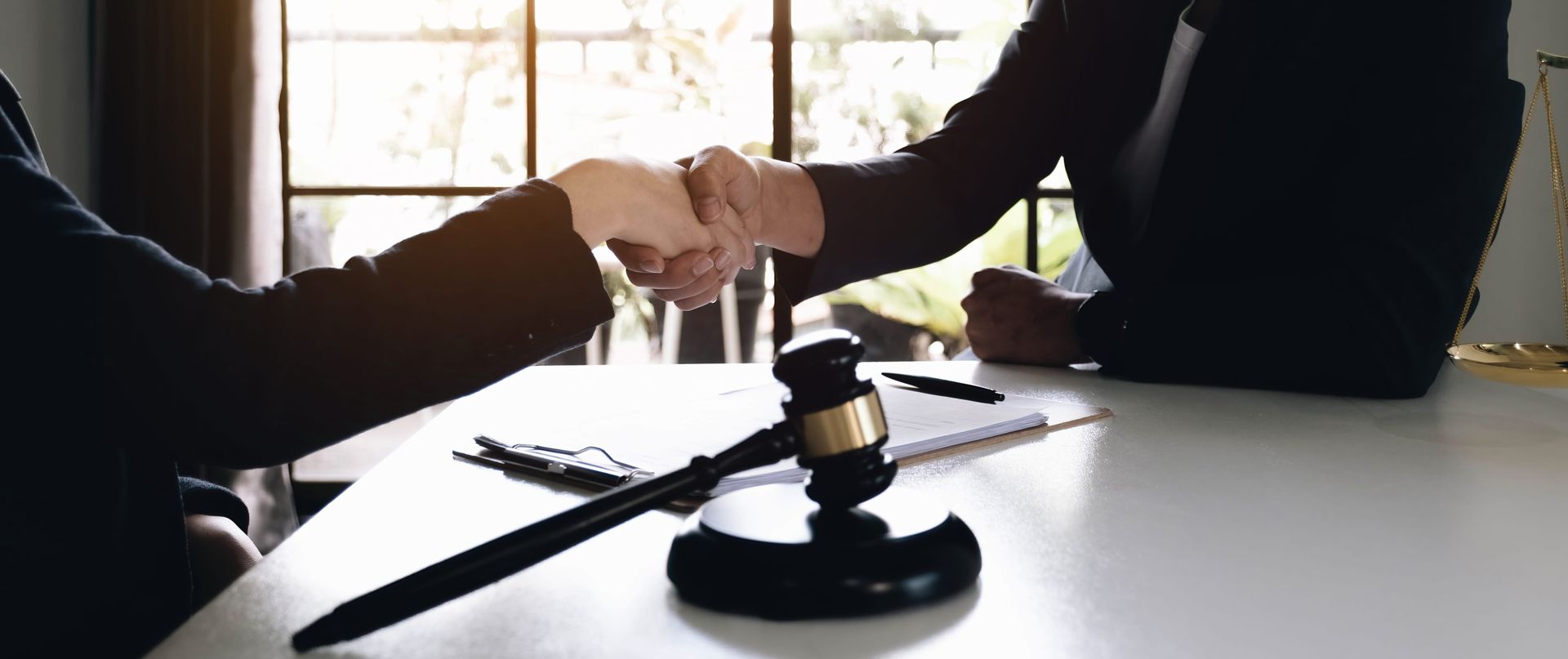 Two people shaking hands over a wooden gavel on a table in a brightly lit office setting.