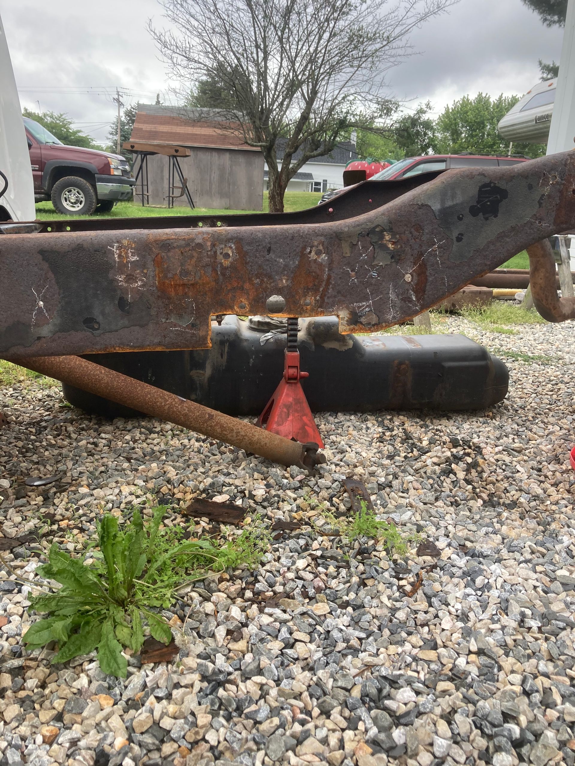 Rusty vehicle frame supported by jack stands; a gas tank and gravel ground.