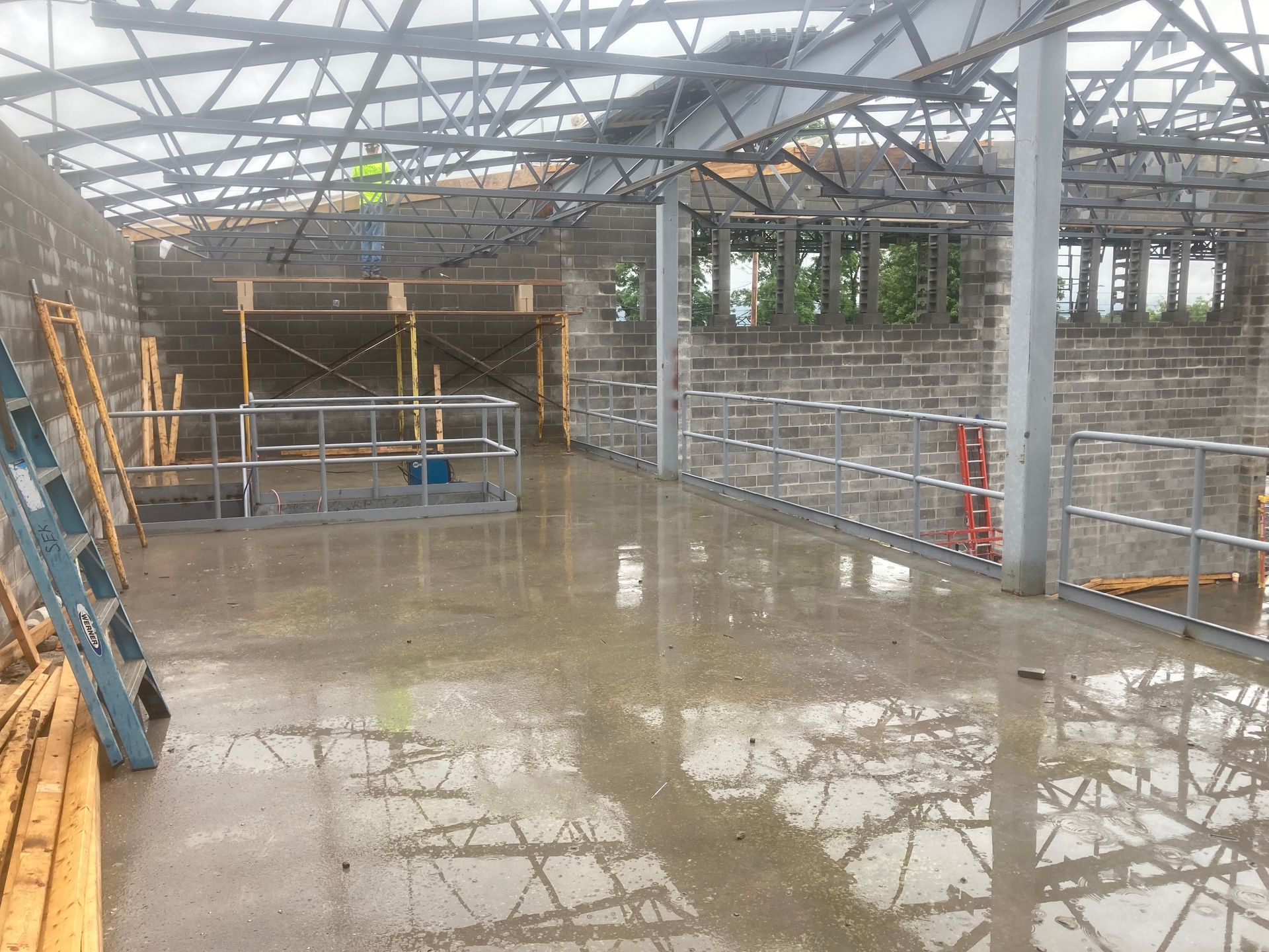 Construction site with wet concrete floor, exposed metal framework, and brick walls. Scaffolding visible.