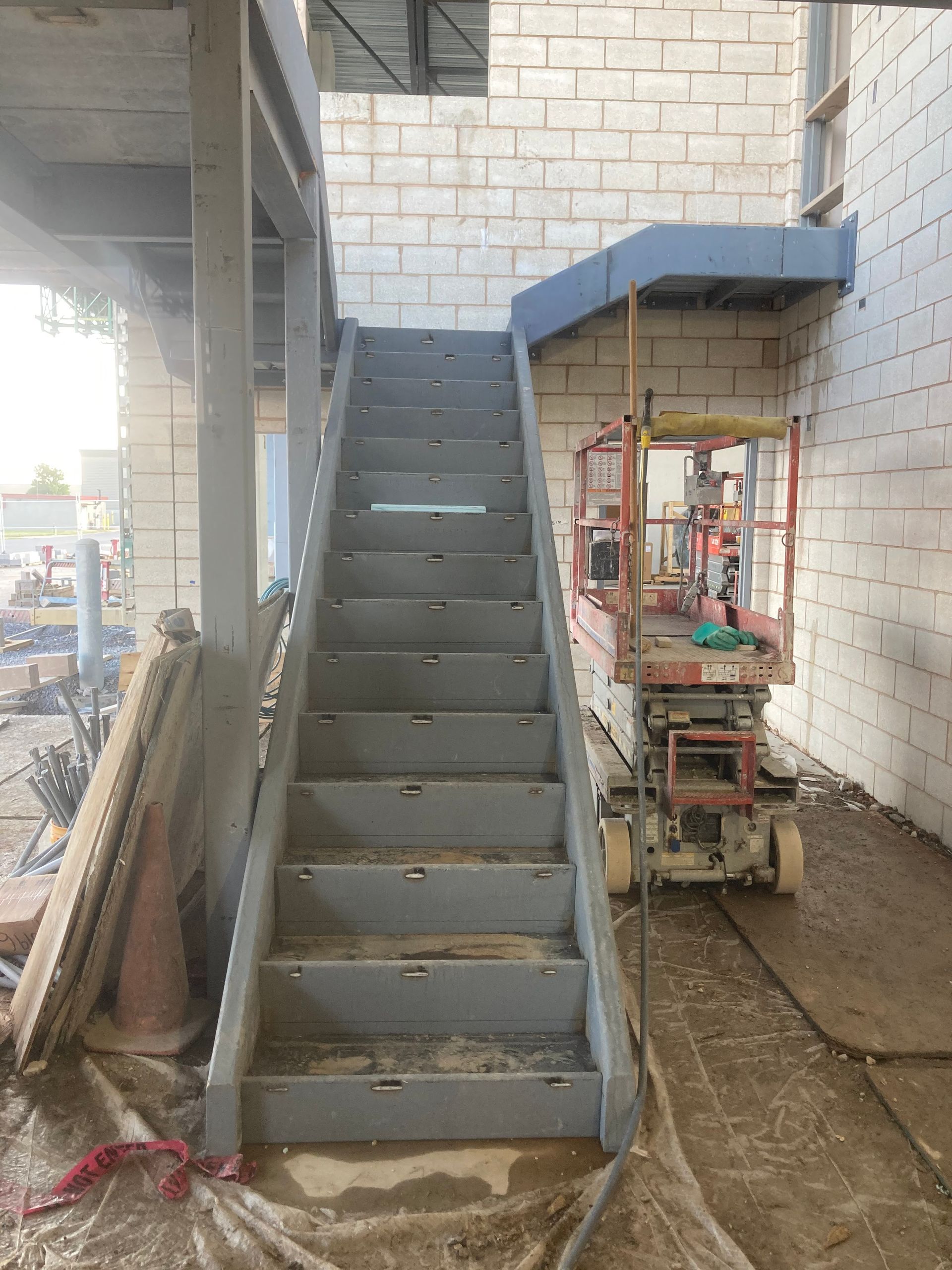 Construction site with gray metal stairs, scaffolding, and a lift. Building has light-colored brick walls.