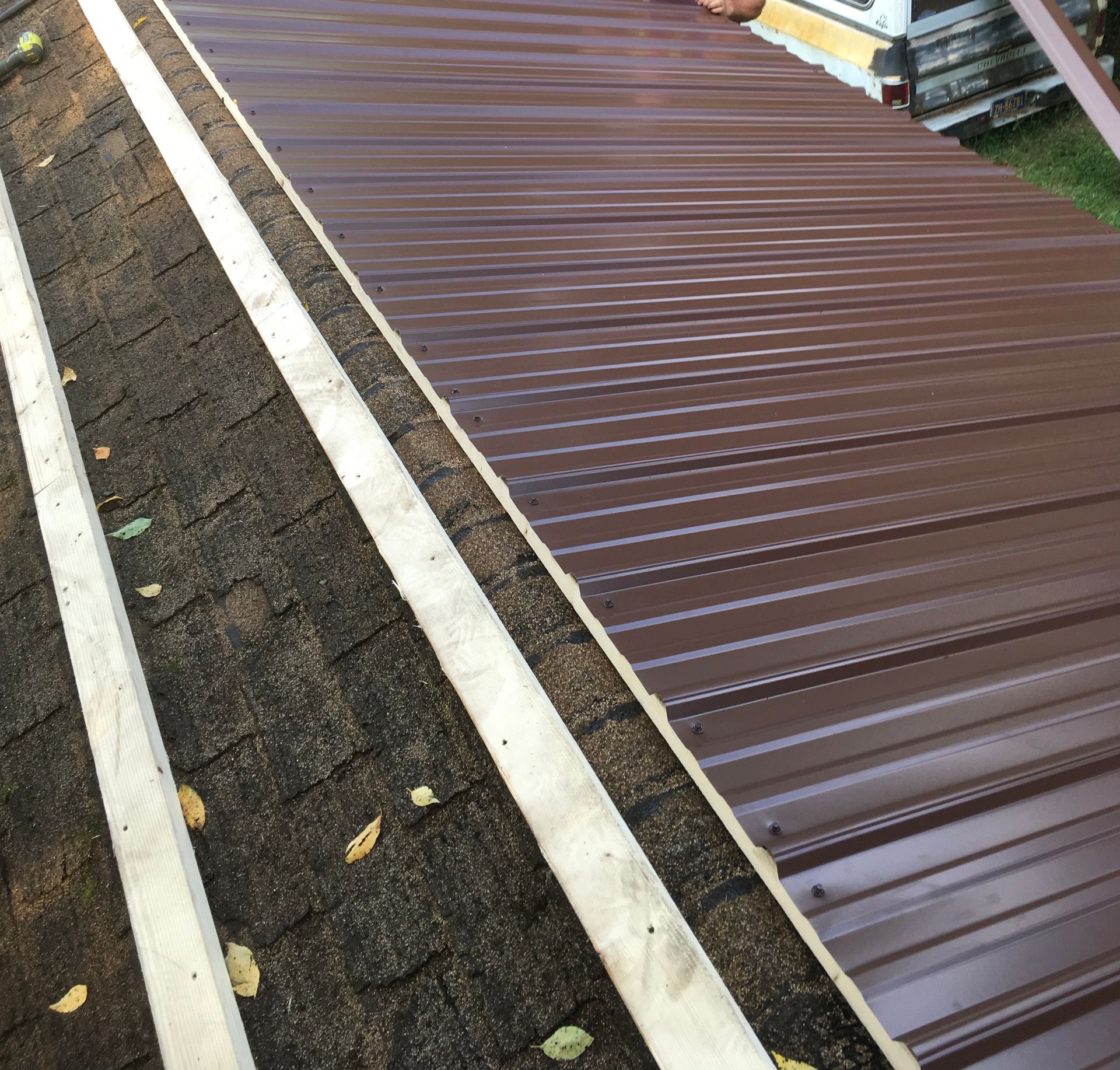 Brown metal roofing being installed over wooden supports on a roof covered in dark shingles.