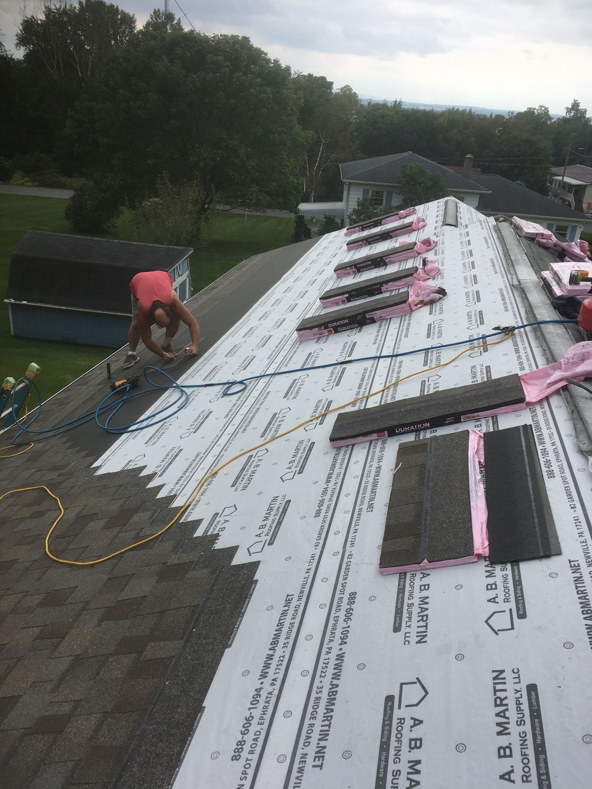 Roofer installing shingles on a roof. White underlayment and pink insulation are visible. Overcast day.