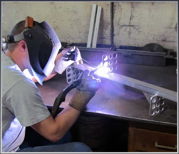 Person welding metal with a mask, sparks flying, in a workshop setting.