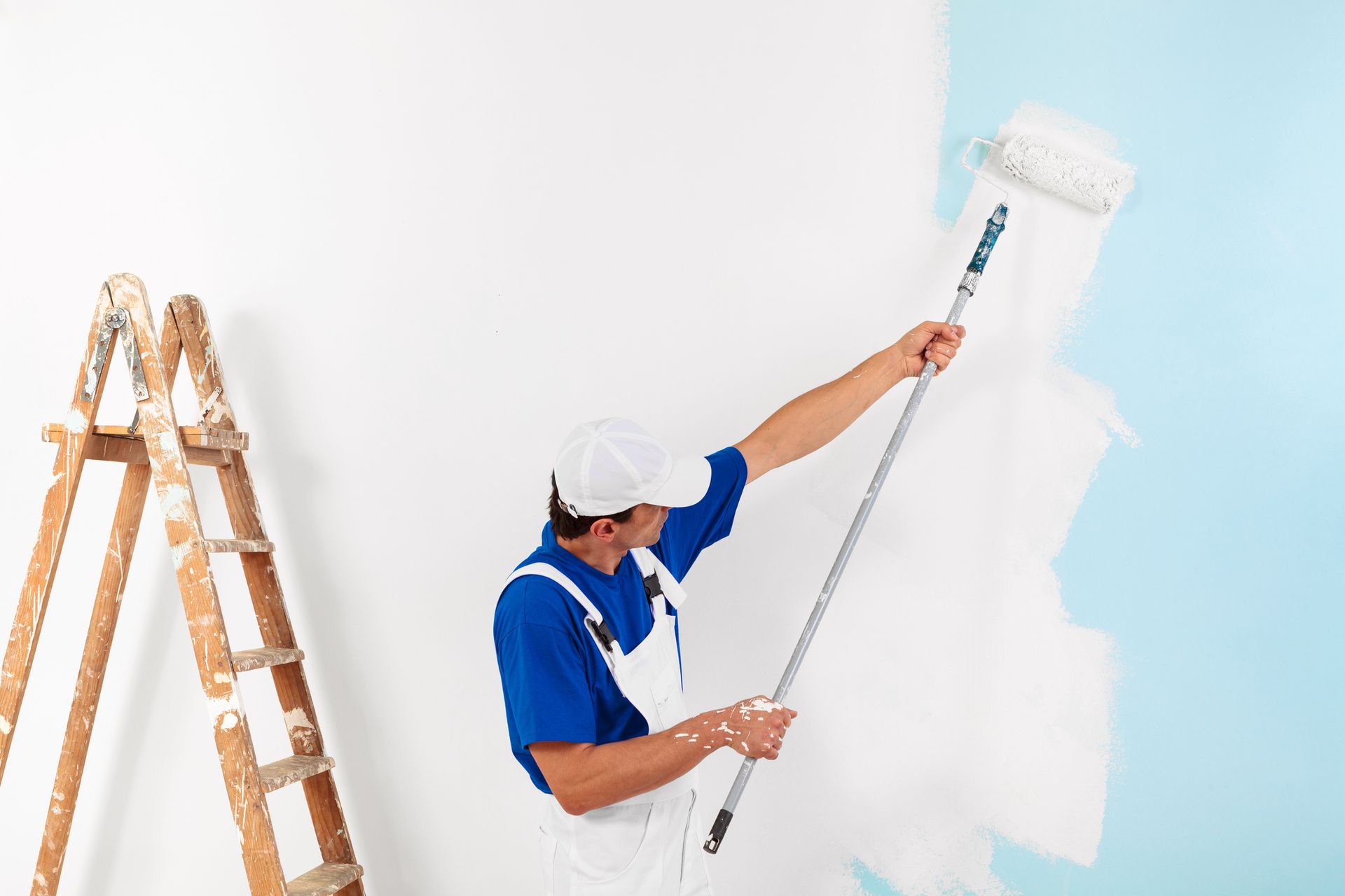 Male painter uses a paint roller to paint a blue wall with white paint, next to a stepladder.