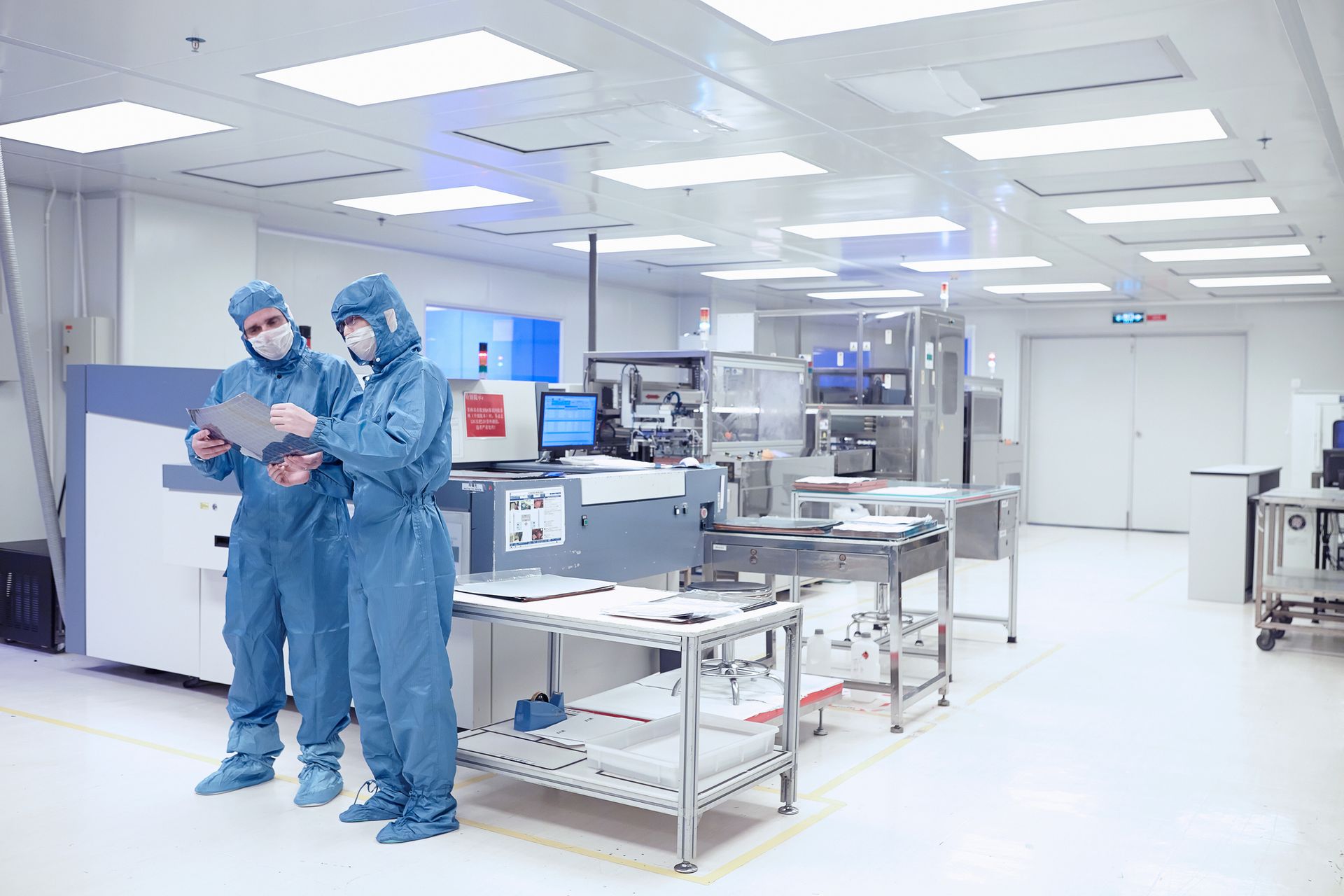 Two people in protective suits are standing in a clean room looking at a tablet.