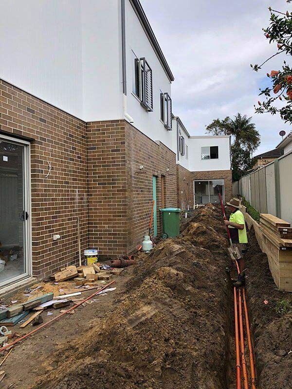 A Man Is Digging A Trench In Front Of A Brick Building — EESI Electrical Contractors In Alstonville, NSW