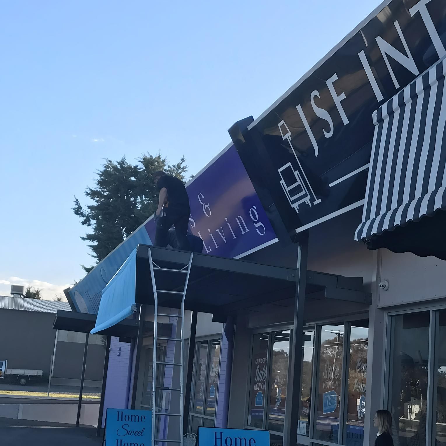 Man On Ladder Repairing Storefront Sign With Black And White Awning — JSF Interiors Goulburn in Goulburn, NSW