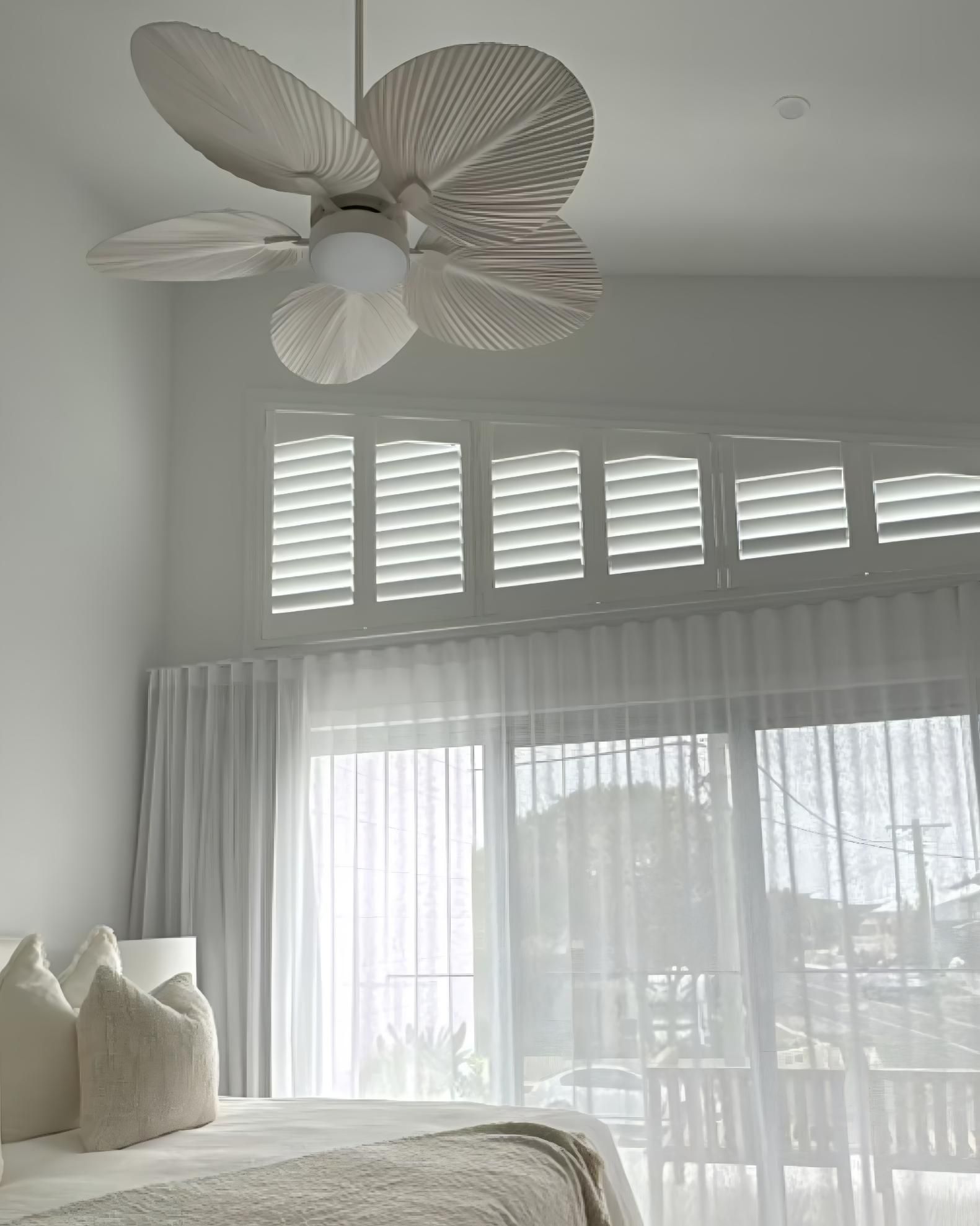 Bedroom with white walls, large windows with shutters and sheer curtains, a bed, and a decorative ceiling fan.
