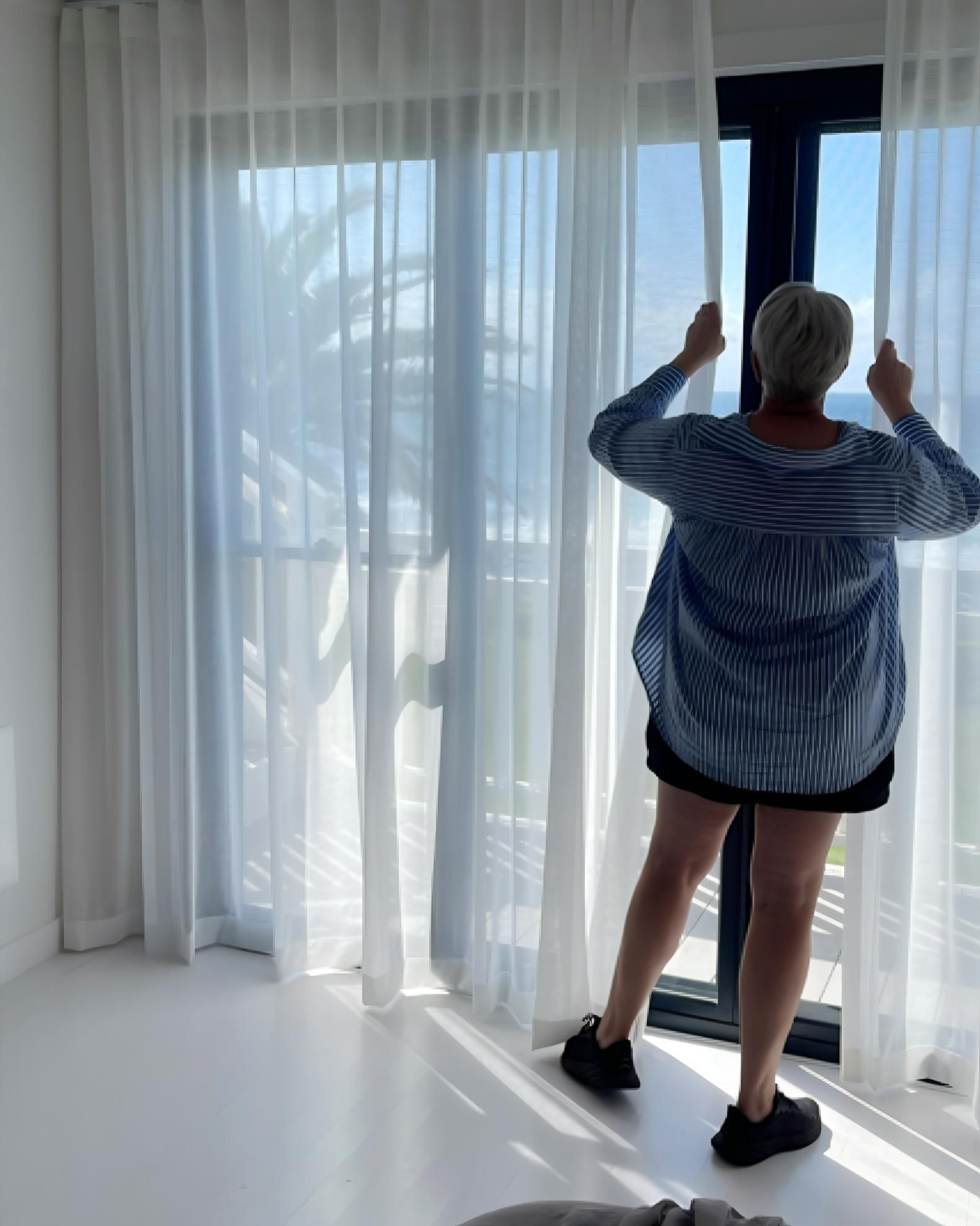 Woman Opening Sheer White Curtains With Ocean View — JSF Interiors Goulburn in Goulburn, NSW