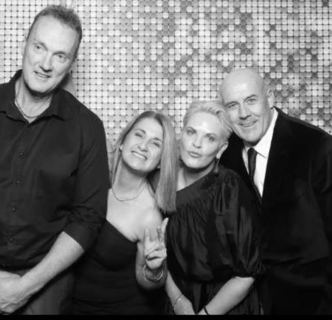 Four people smiling in front of a sequined backdrop, black and white photo — JSF Interiors Goulburn in Goulburn, NSW