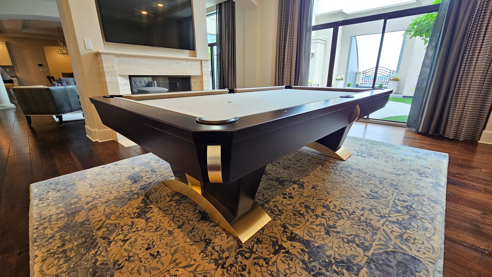 Pool table in a luxury living room. Dark wood, gold accents, light rug, fireplace, and large window.