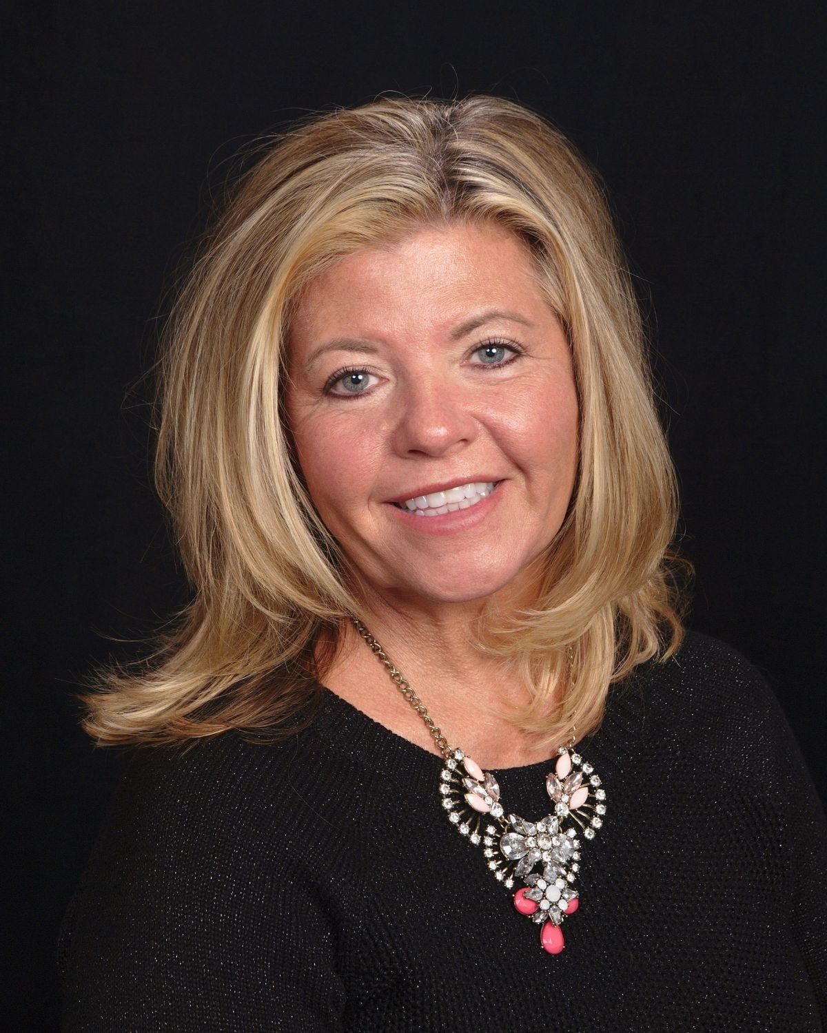 Smiling woman with blonde hair, wearing a black top and necklace, against a black background.
