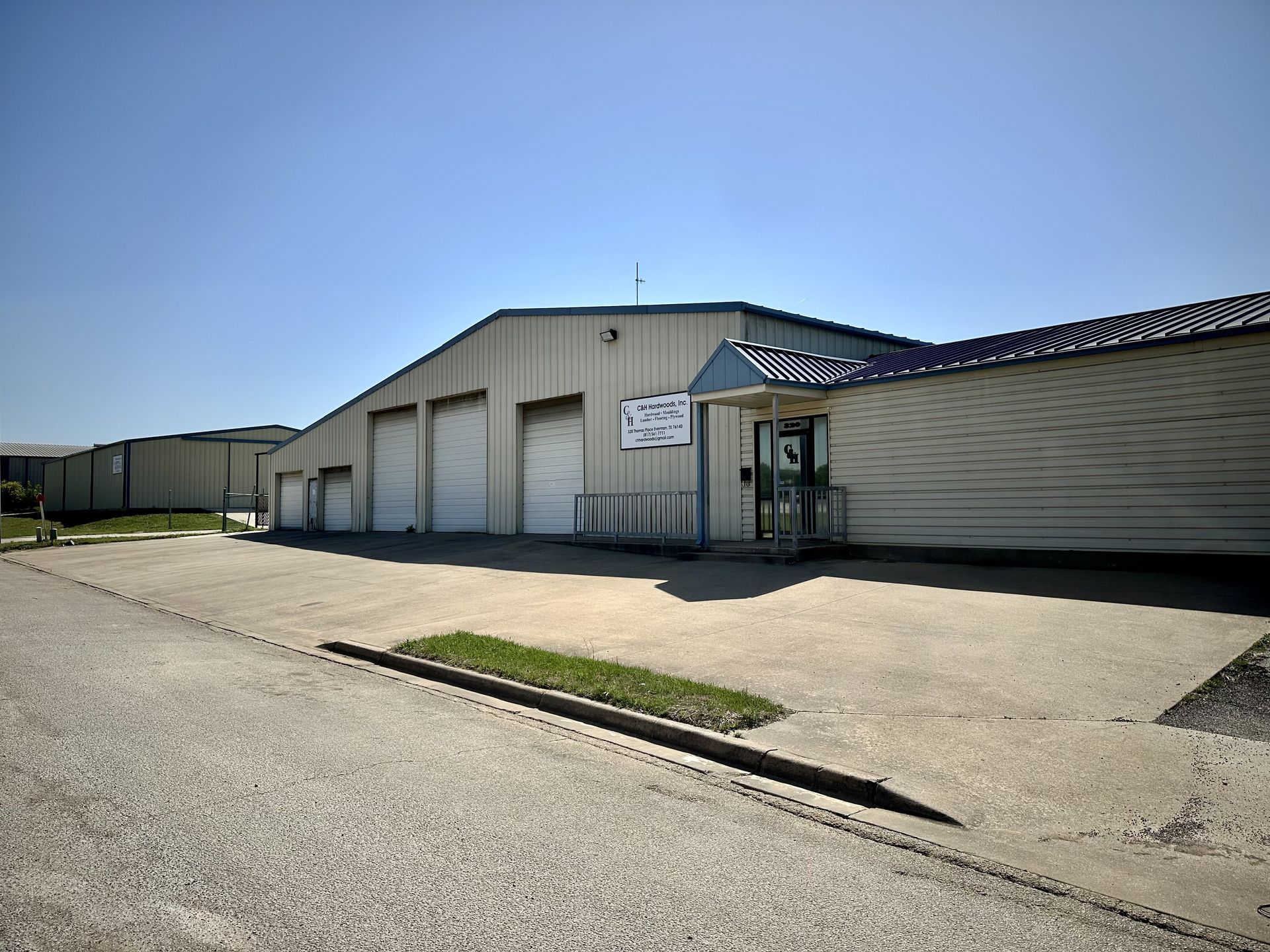 A commercial building with three garage doors and an entrance under a blue awning. Bright sunlight casts shadows on the asphalt.
