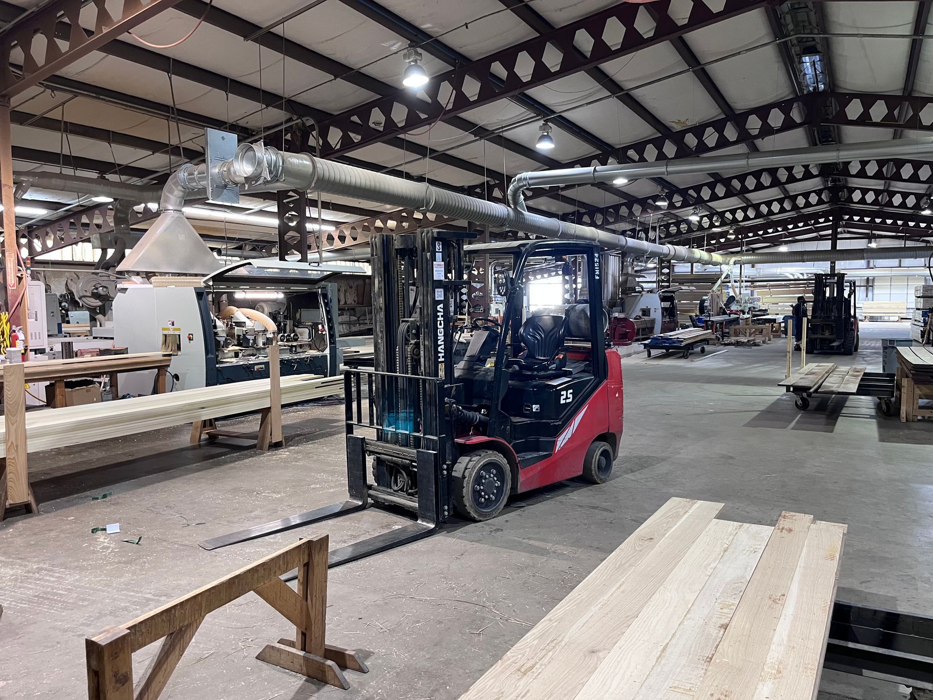 Forklift operating in a wood workshop, moving lumber. The workshop has industrial equipment, exposed beams, and a ventilation system.
