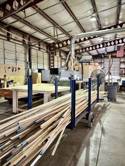 A woodshop interior with stacks of lumber on a cart, machinery, and tools, ready for work. An American flag hangs on a wall.