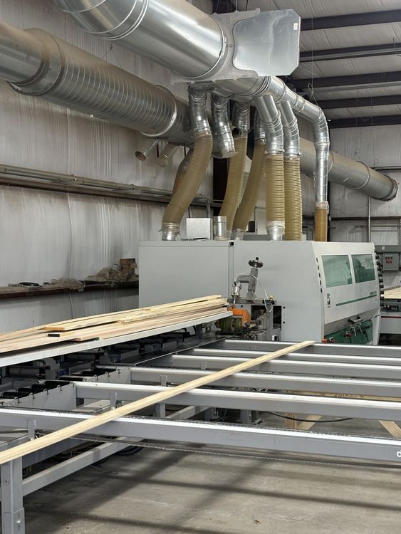 Factory machine processing wood, with ventilation system above.  Long wooden planks are moved along a metal track in a workshop.