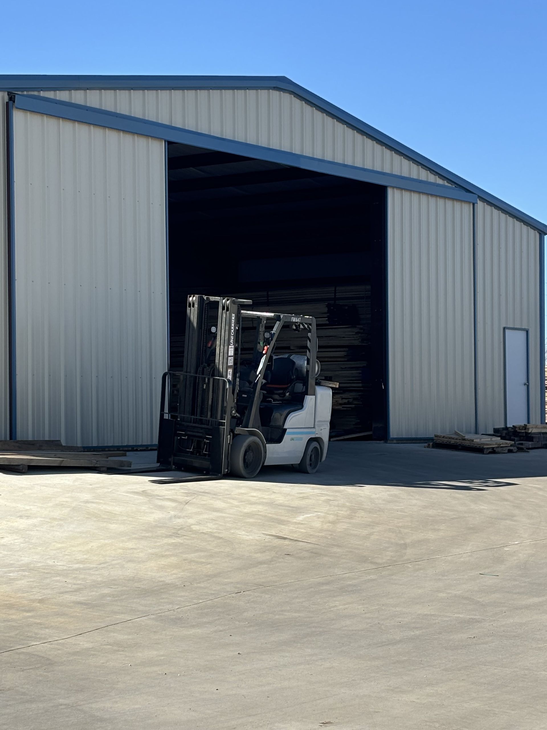 A white forklift exits a large warehouse with a wide open door. The building is light-colored with a blue roof.