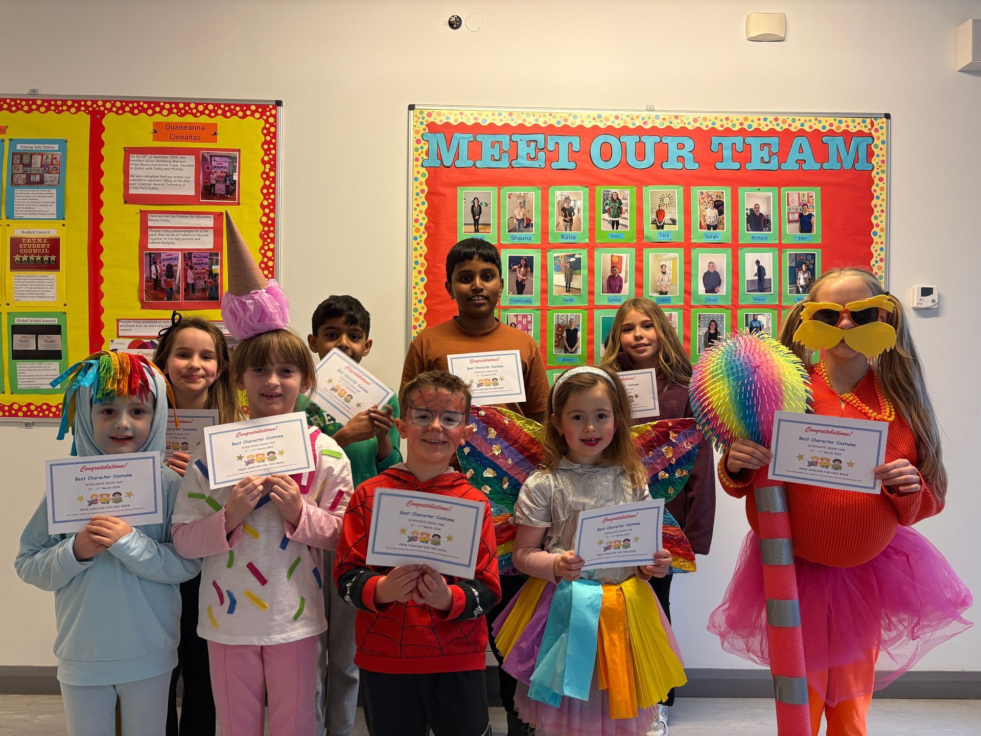 Children holding certificates pose in a classroom decorated with colorful bulletin boards and a “MEET OUR TEAM” display.