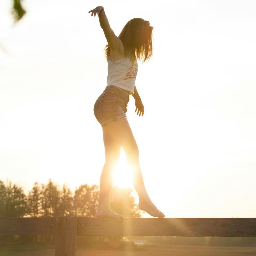 Woman balancing on a wooden fence, silhouetted against a bright, sunny sky.