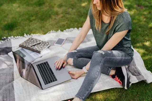 A woman is sitting on a blanket in the grass using a laptop computer.