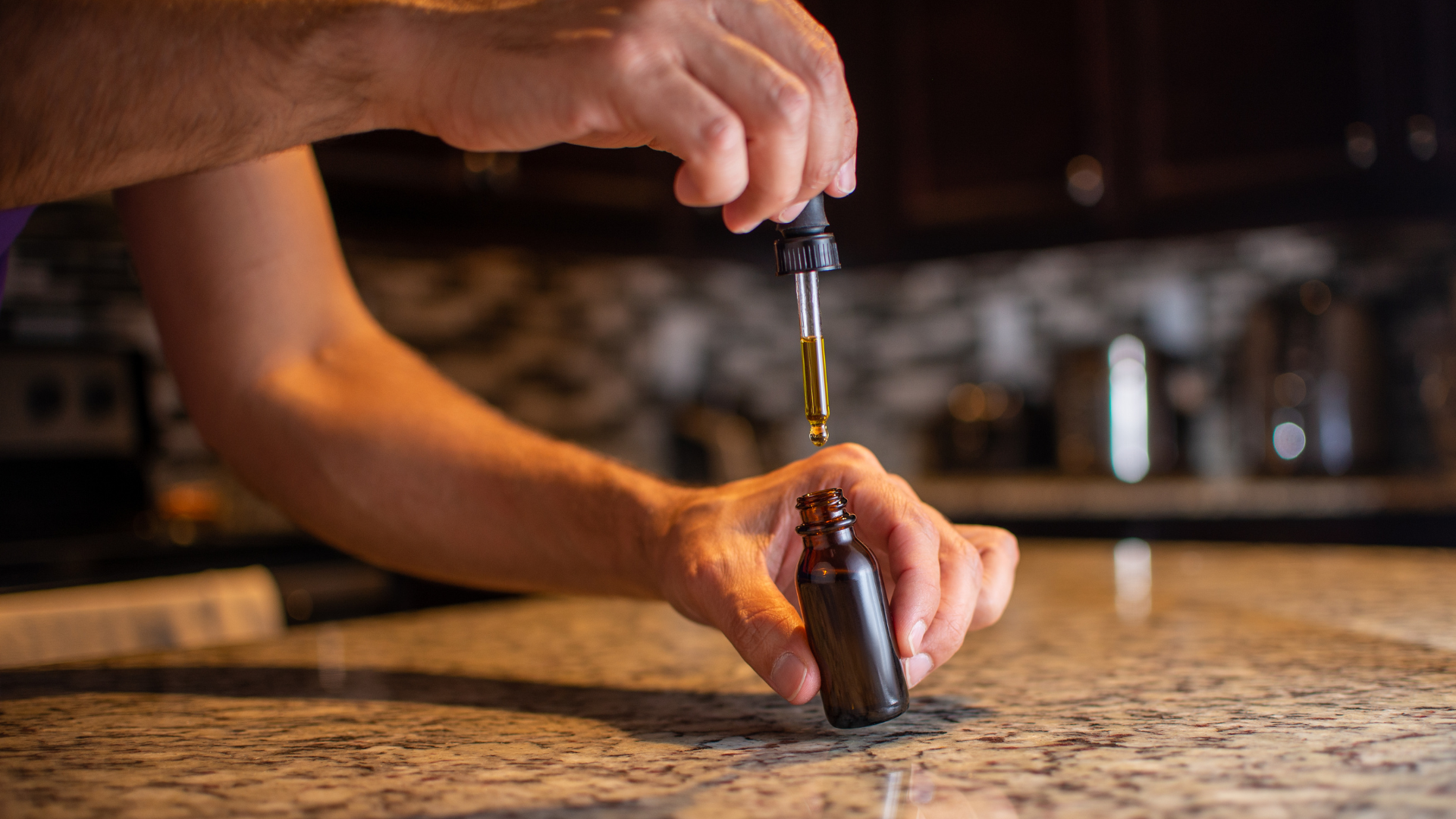 Person dispensing oil from a dropper onto their wrist, bottle in hand, in a kitchen.