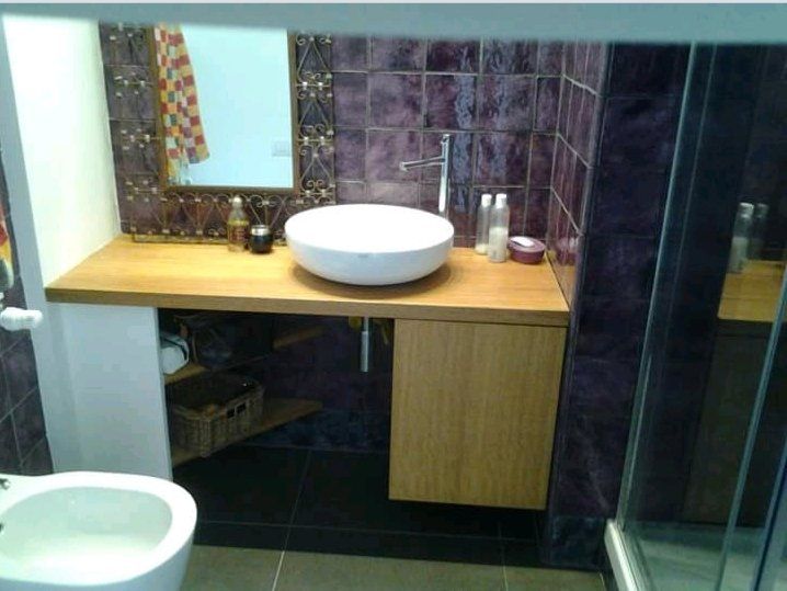 Bagno con lavandino bianco su un piano in legno, parete piastrellata viola e specchio.
