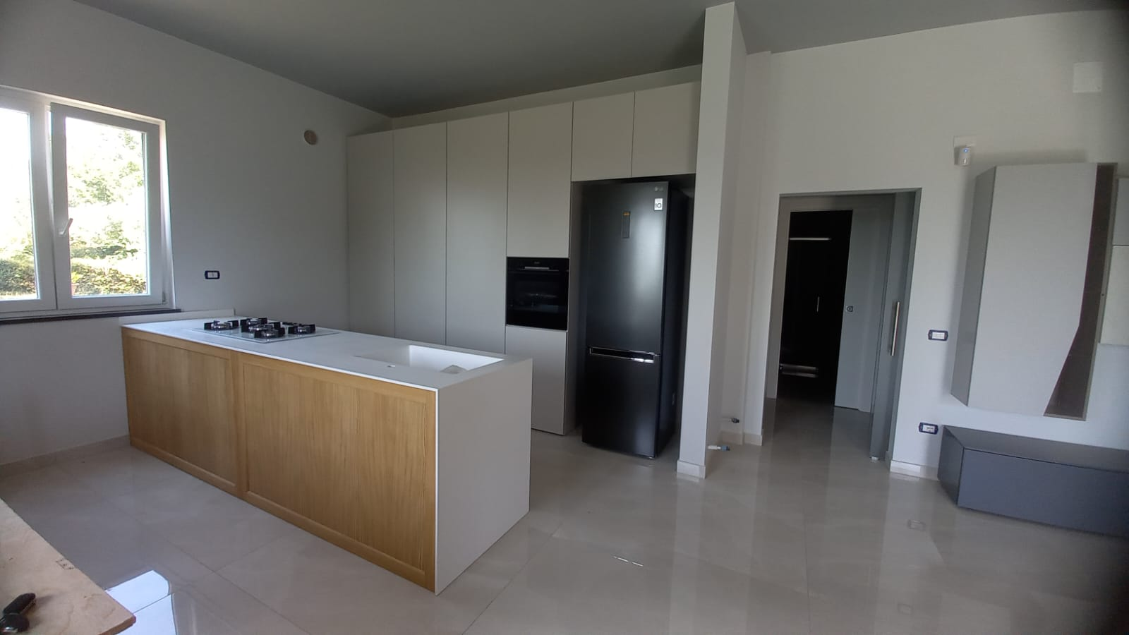 Cucina moderna con isola in legno chiaro, mobili grigi, frigorifero nero e porta aperta.