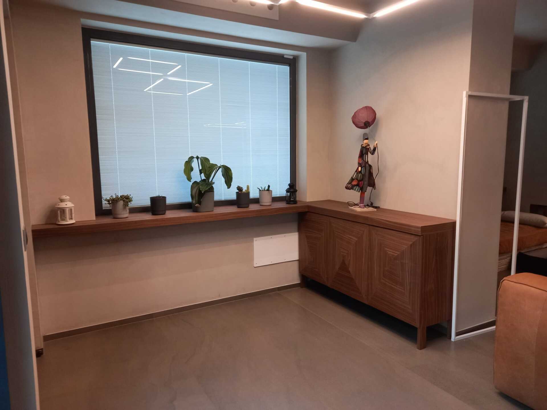 Stanza con finestra, scaffale in legno e mobiletto. Piante verdi sullo scaffale, statuetta sul mobiletto.