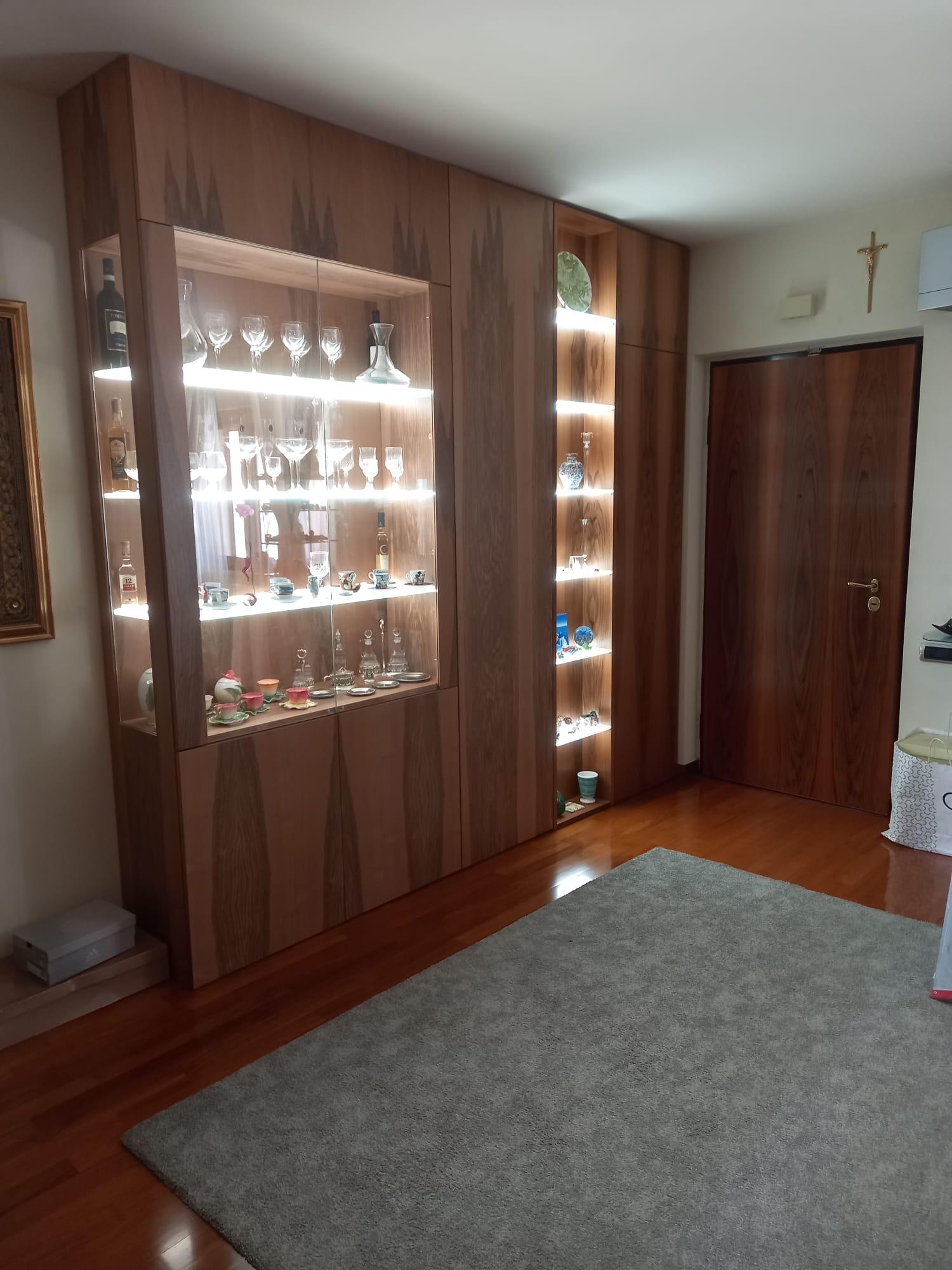 Vetrina in legno con ripiani illuminati contenenti oggetti in vetro, in una stanza con un tappeto e pavimenti in legno.